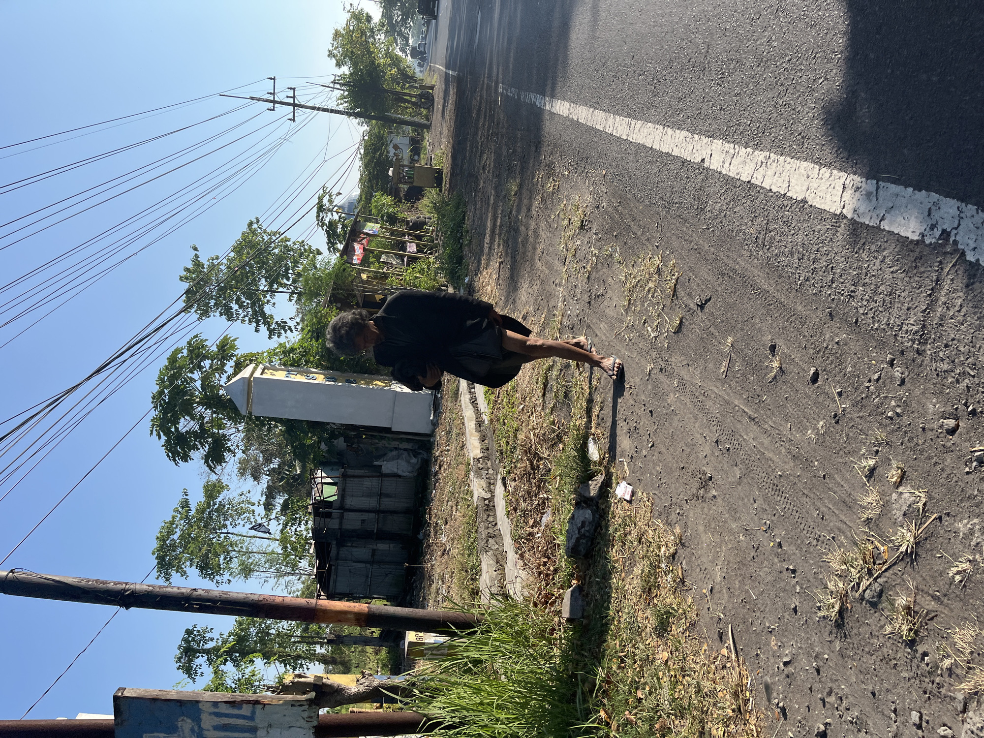 In Siring, Indonesia, a person with unkempt dark hair wearing glasses, a black jacket, and shorts walks on a dirt shoulder beside a paved road. The person is holding a dark item. Behind them, there are utility poles with numerous wires, a building with corrugated metal siding, and a white pillar. Vegetation, including tall grass and trees, is visible. The road has a white line marking its edge. In the distance, vehicles are on the road, and more trees line the sides. The sky is clear and blue.