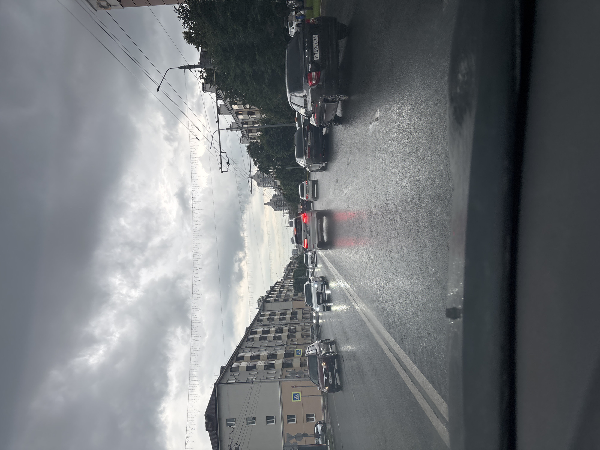 The image depicts a street scene in Kazan, Russia, likely during daylight hours. The sky is heavily overcast with dark, dramatic clouds, suggesting a gloomy or stormy weather condition. The road is wet, indicating recent rain or ongoing precipitation, with cars driving in a line, suggesting a traffic jam or slow-moving traffic.
Prominently featured are several cars of various models, their headlights and taillights visible. The cars are driving on the right side of the road. On the left side of the street, there are multi-story apartment buildings with a uniform architectural style, typical of Soviet-era housing. Trees are visible along the sidewalk and between buildings. Overhead, there are power lines and streetlights, along with decorative string lights suspended across the street, possibly for an event or holiday.
One car in the foreground, a black Lada, has a license plate that appears to read "C 7699043". Another car further down the road also has a visible license plate. Pedestrian crossing signs are also visible on the sidewalk. The overall mood is somewhat somber due to the weather and the traffic congestion. The perspective is from inside a vehicle, looking out at the scene.