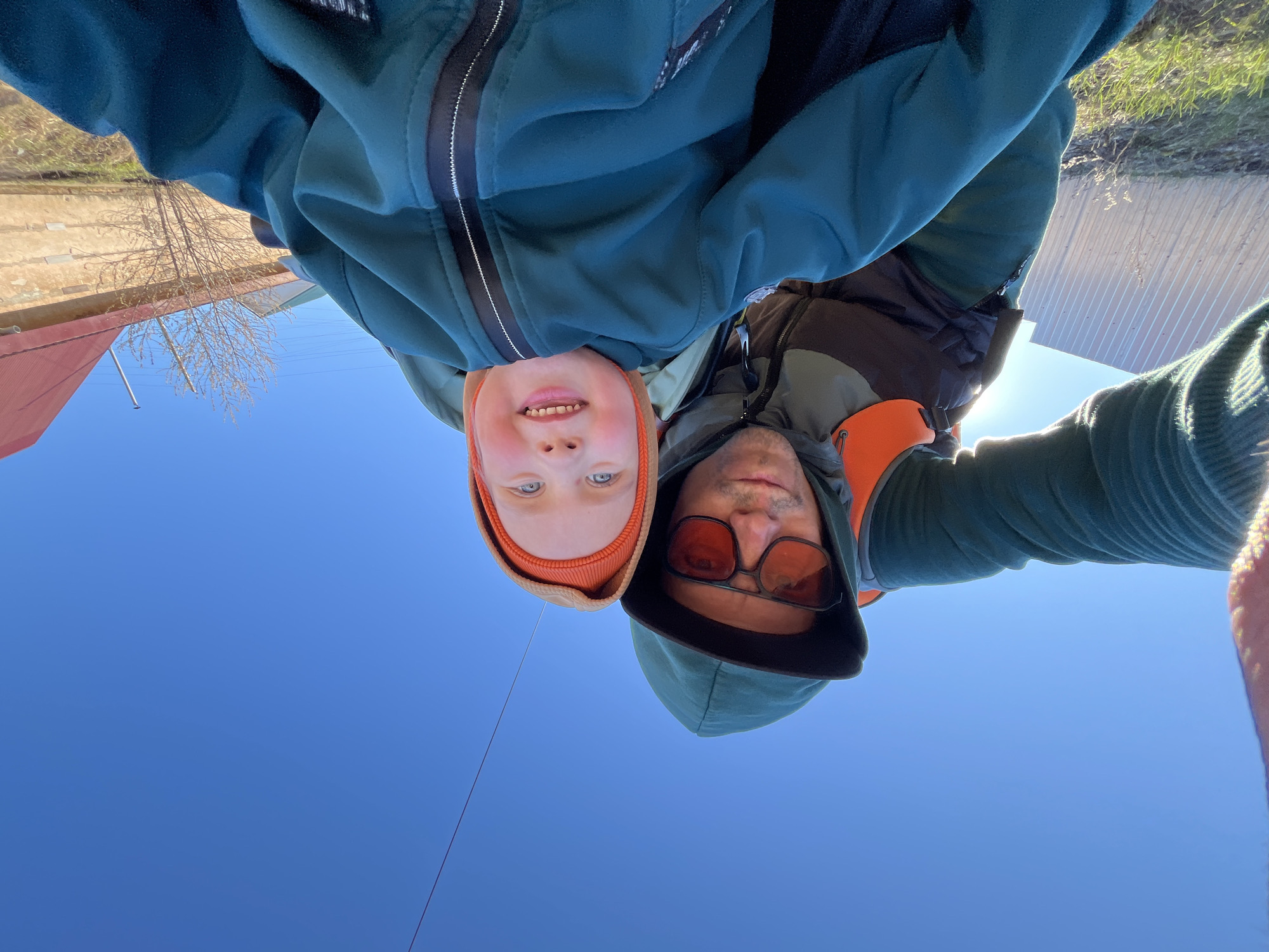 A selfie-style, upside-down angle photograph shows a fair-skinned young child and an adult, likely male, in outdoor attire against a backdrop of a clear blue sky and a cityscape reflection.
The child is positioned on the left, wearing an orange knit hat, and smiling with visible front teeth. The adult is on the right, sporting sunglasses with orange lenses and a hooded jacket.
The reflection on the dark surface beneath them suggests the location is near a city street with buildings, possibly in Chaikovsky, Russia. A red roof is visible on the left and a metal surface is visible on the right of the frame. The photo has a single dark, thin, diagonal line running through the middle.