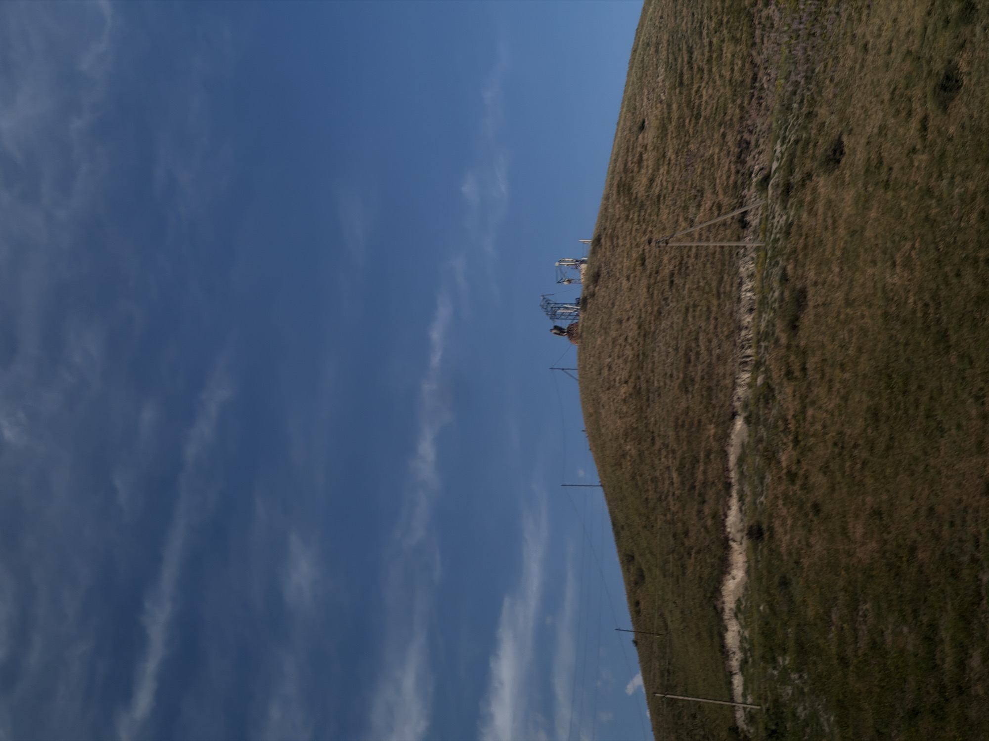 This media file captures a sunlit outdoor scene, possibly taken on a clear day with a few wispy clouds. The foreground is dominated by a grassy hillside, sloping upwards towards the right of the frame. The grass appears dry and brownish, suggesting a warm season. A faint path or track can be seen winding its way up the hill.
In the mid-ground, a series of tall, dark poles with wires strung between them are visible, indicating the presence of a power line or some other form of overhead infrastructure. Further up the slope, there are some complex structures that resemble industrial equipment, possibly related to mining or some form of extraction, with metallic frameworks and what might be cranes. These structures are silhouetted against the bright blue sky.
The sky itself takes up a significant portion of the left side of the image and is a vibrant blue, streaked with thin, white clouds. The overall impression is one of a vast, open landscape under a bright sky. There is no visible text in the image. The scene appears to be remote and undeveloped, with the natural landscape being the primary feature, interspersed with man-made structures.