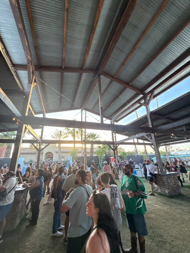 The image captures a scene at what appears to be an outdoor event, likely a music festival or similar gathering, in Indio, United States. A large open-air structure with a corrugated metal roof supported by wooden beams covers the area. A crowd of people is gathered beneath the structure, suggesting a place to relax, socialize, and access refreshments. Attendees are dressed in various casual styles. Some are standing at stone-built bar counters, engaging in conversation and likely ordering drinks. Palm trees and buildings can be seen in the background, bathed in the warm light of the setting sun.