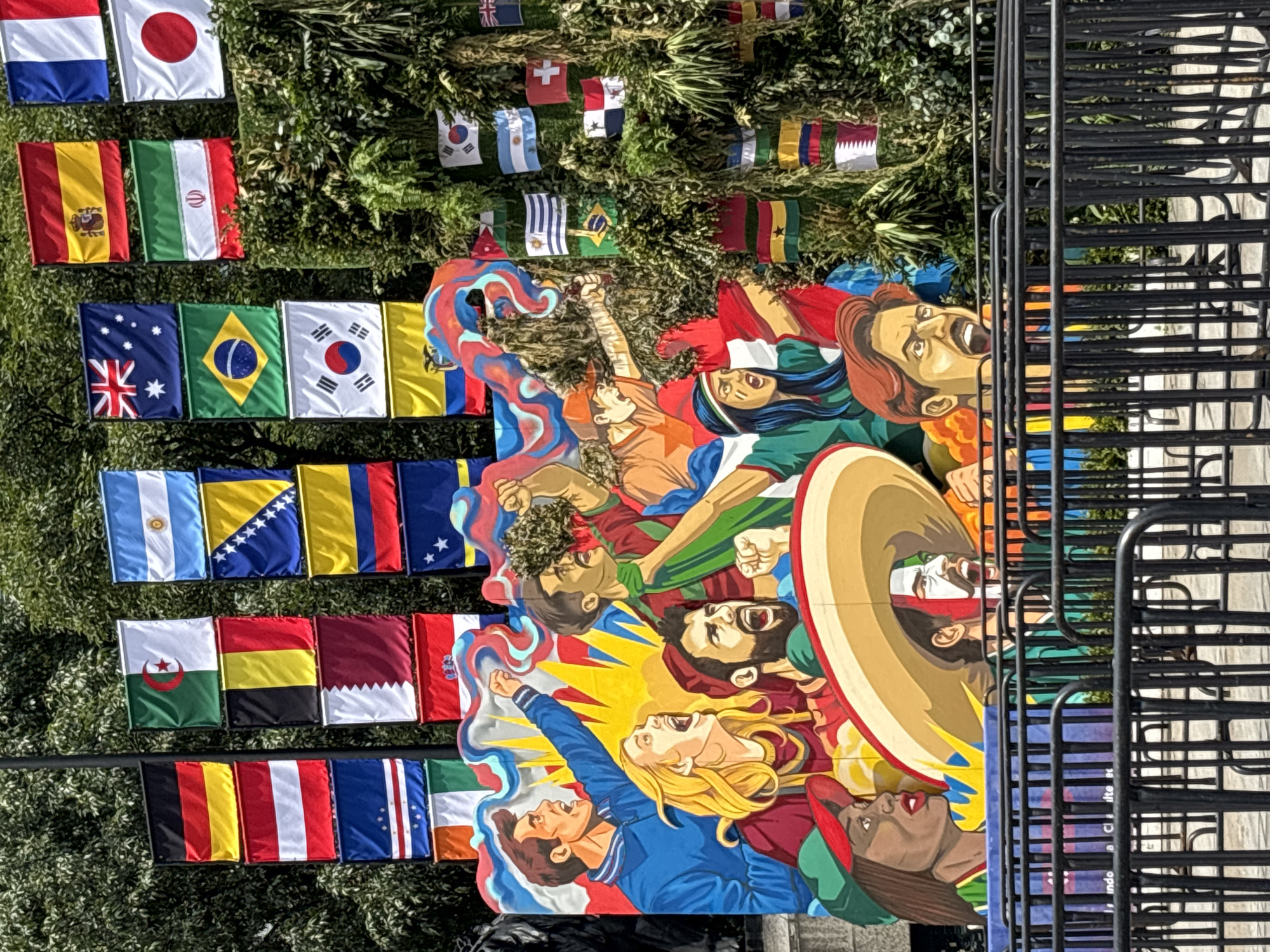 The image depicts an outdoor public art installation in Mexico City, Mexico. The installation features a colorful mural of diverse people depicted with intense expressions, many with their mouths open as if cheering or shouting, suggesting a theme of celebration or strong emotion. Surrounding the mural and integrated into a densely planted green structure are numerous national flags, indicating a possible international or sporting event theme.
The mural's foreground shows several figures rendered in a stylized, almost cartoonish manner. One individual wears a large sombrero and has their face painted with red and white stripes. Another figure has dark skin and wears a green cap. Behind them, a group of individuals, primarily depicted with lighter skin tones, are shown in various states of vocalization, with one in a blue jacket raising a fist. The artistic style is bold and graphic, with sharp lines and vibrant colors.
The backdrop consists of a dense arrangement of national flags of various countries, including but not limited to Germany, Argentina, Spain, Brazil, Australia, South Korea, and Mexico. These flags are hung in a vertical cascade and also appear integrated into the greenery. The foliage itself is lush and textured, adding a natural element to the otherwise man-made display.
In the foreground, black metal barricades are set up, suggesting the area is cordoned off for public viewing or an event. The ground behind the barricades is paved. The overall impression is a lively and patriotic display, likely related to a significant cultural or sporting occasion.