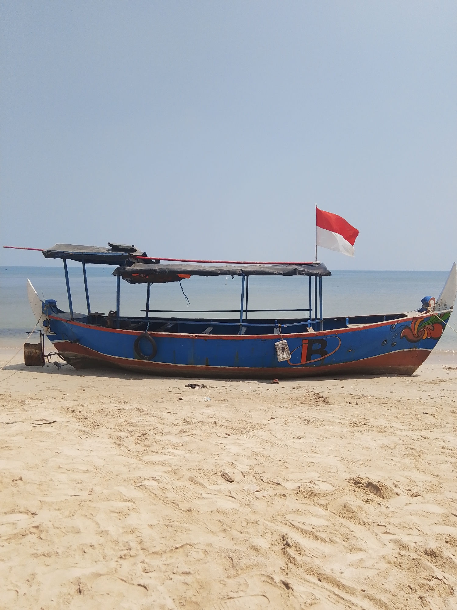 An Indonesian flag, red and white, flies from a pole on a blue and red wooden boat. The boat is beached on a sandy shore. The sea is calm and blue, meeting a clear, pale blue sky. The boat has a canvas roof supported by poles. The hull is painted blue with a red lower section. A white decorative flourish is visible on the stern. The sand is textured with tracks and small debris. There is no visible human activity.