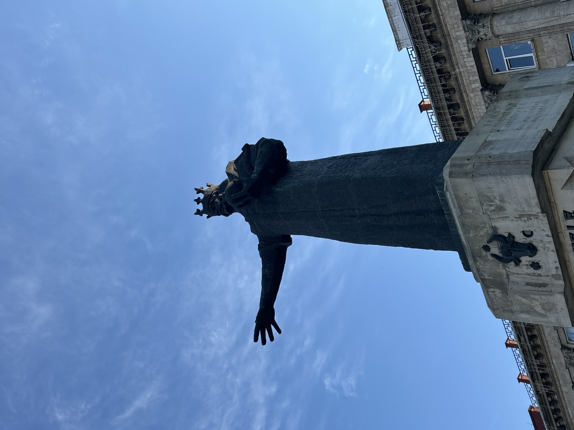 The image shows a low-angle shot of the Stephen the Great monument in Bălți, Moldova, against a backdrop of a bright blue sky with sparse white clouds. The dark bronze statue of Stephen the Great, wearing a crown, stands tall on a stone pedestal. His right arm is extended outwards, palm open, in a gesture of welcome or command. The pedestal features inscriptions and decorative elements, including a bull's head motif. Behind the monument, the upper floors of a classical-style building are visible, characterized by ornate architectural details, columns, and windows. A line of decorative lights or garlands is strung across the building's facade. The overall scene captures a significant historical landmark in the city.