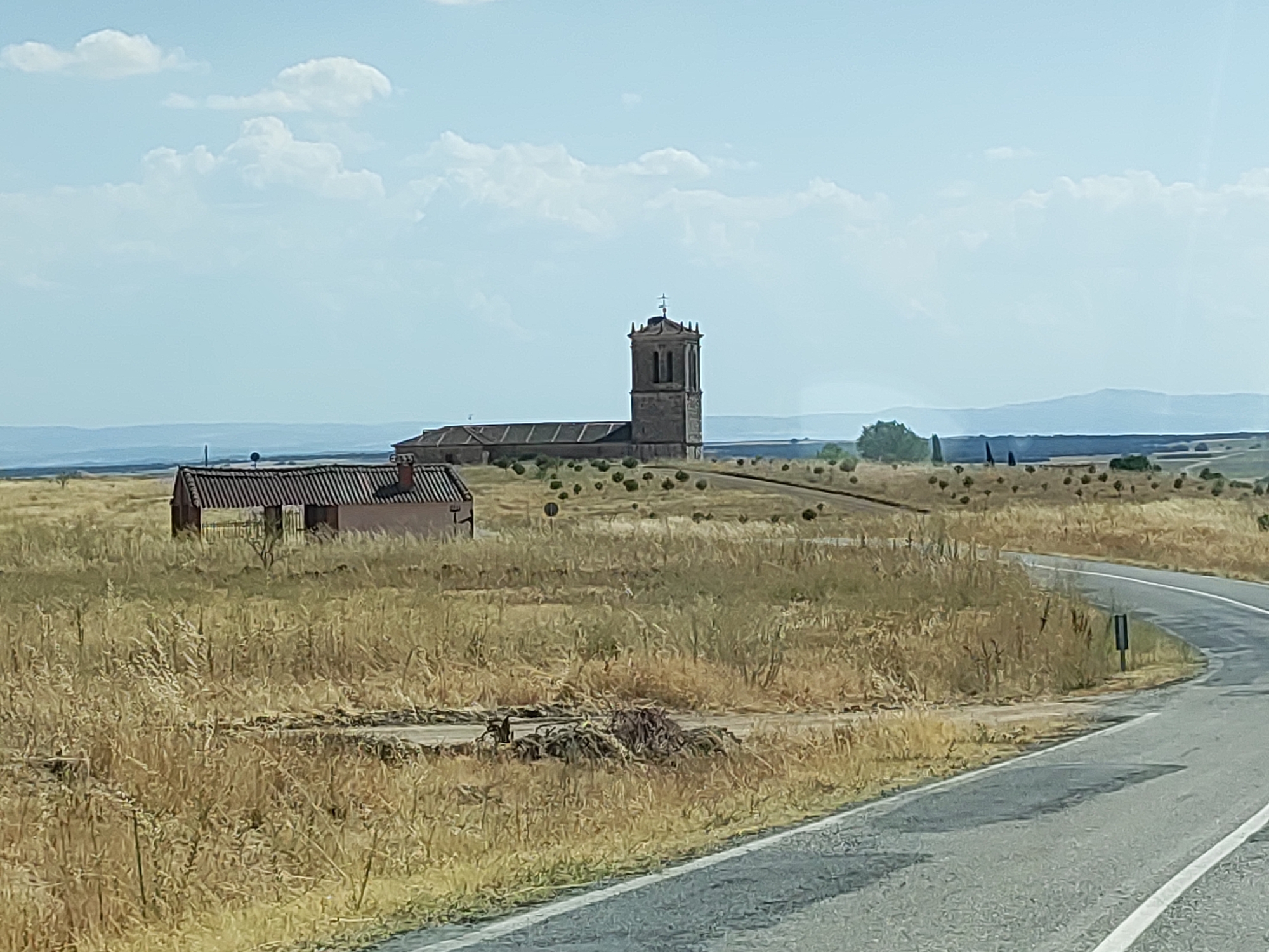 The image depicts a rural landscape with a church and a small building in the distance. A paved road curves through the foreground, bordered by dry, golden grass. The sky is partly cloudy with a pale blue hue. The church features a prominent bell tower with a cross at its apex and a long, rectangular nave. A small, single-story building with a tiled roof and a chimney is situated to the left of the church. The surrounding terrain is characterized by gently rolling hills dotted with sparse vegetation and a few trees. The scene appears to be bathed in daylight. The location is identified as Santa María la Real de Nieva, Spain.