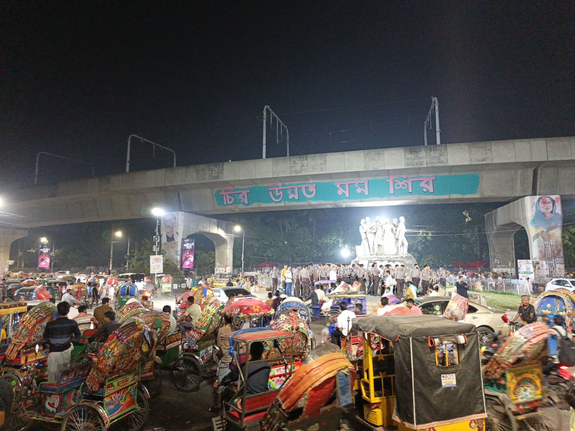 The image depicts a busy intersection in Dhaka, Bangladesh at night, identifiable by the presence of numerous cycle rickshaws, commonly used as transportation in the city. The rickshaws are brightly decorated with colorful designs. Above the intersection is an overpass with Bengali text written on it, which, coupled with the banners featuring Bengali script and political figures, places the location firmly in Bangladesh.
A group of uniformed police officers are gathered near a white monument, likely a memorial or public art installation. The environment appears urban with streetlights illuminating the scene. There are cars and other vehicles amidst the rickshaws, suggesting heavy traffic. The overall scene is one of bustling urban activity in Dhaka.