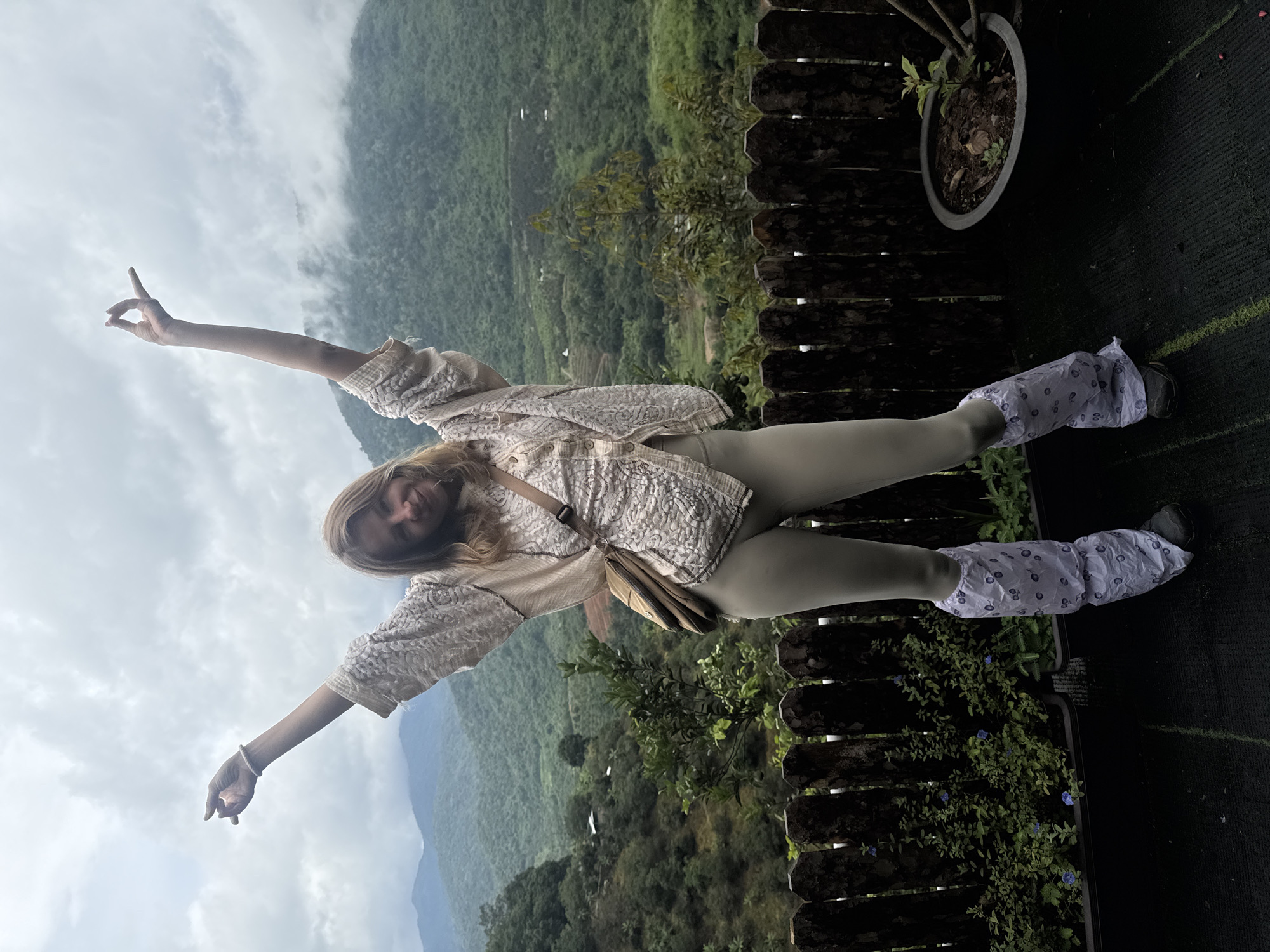 A young woman stands on a dark, textured platform in front of a wooden fence. She is wearing a light-colored, patterned blouse over a light-colored top, light-colored leggings, and leg warmers with a floral pattern. She has her arms raised, with her right hand pointing upwards and her left hand in a gesture with her thumb and index finger touching. She is looking at the camera and smiling. Behind her, lush green mountains covered in clouds stretch into the distance. The platform appears to be an outdoor space, possibly a balcony or viewing area, with plants in dark containers to the left and right of the woman. The sky is overcast. The location is identified as La Da, Vietnam, a mountainous region known for its scenic beauty.
