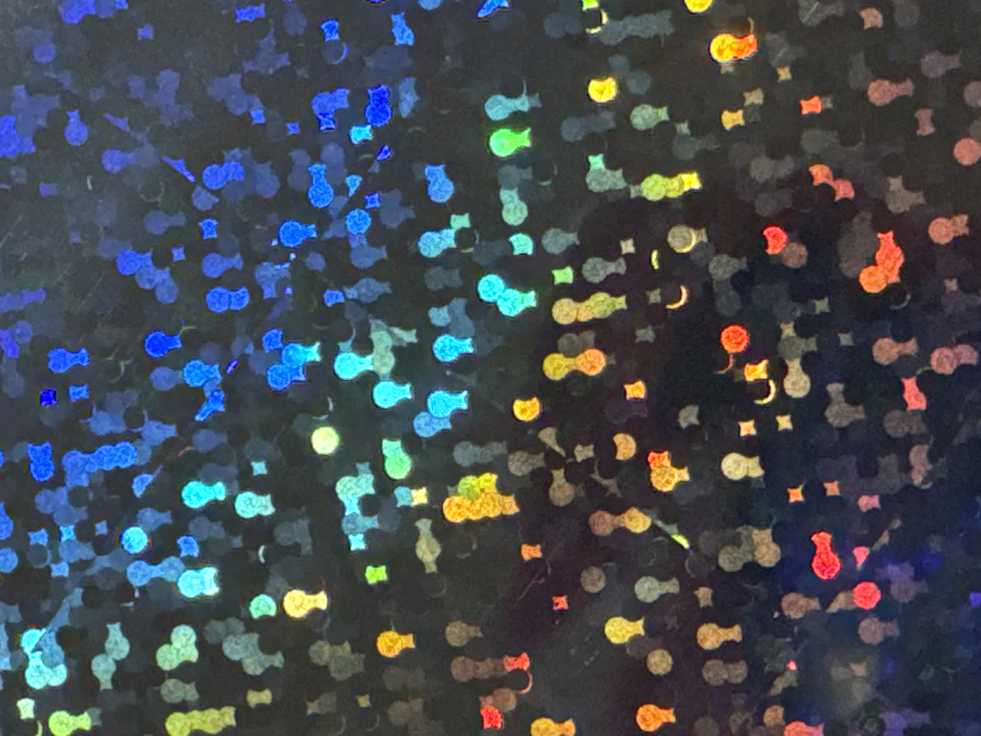 The image is a close-up, abstract shot of a holographic material. It displays a dark background with a multitude of small, reflective shapes in various colors, including blue, green, yellow, orange, and red. The shapes are varied, some appearing as circles, others as elongated forms with rounded tops resembling tiny upside-down lightbulbs, and some as squares. The holographic effect creates a shimmering, iridescent quality, with colors shifting and blending. The lighting is diffuse, highlighting the reflective nature of the material. There are no discernible people, objects, or environmental features that would suggest a specific location within the city of Waterford, Ireland. The image is purely textural and chromatic.