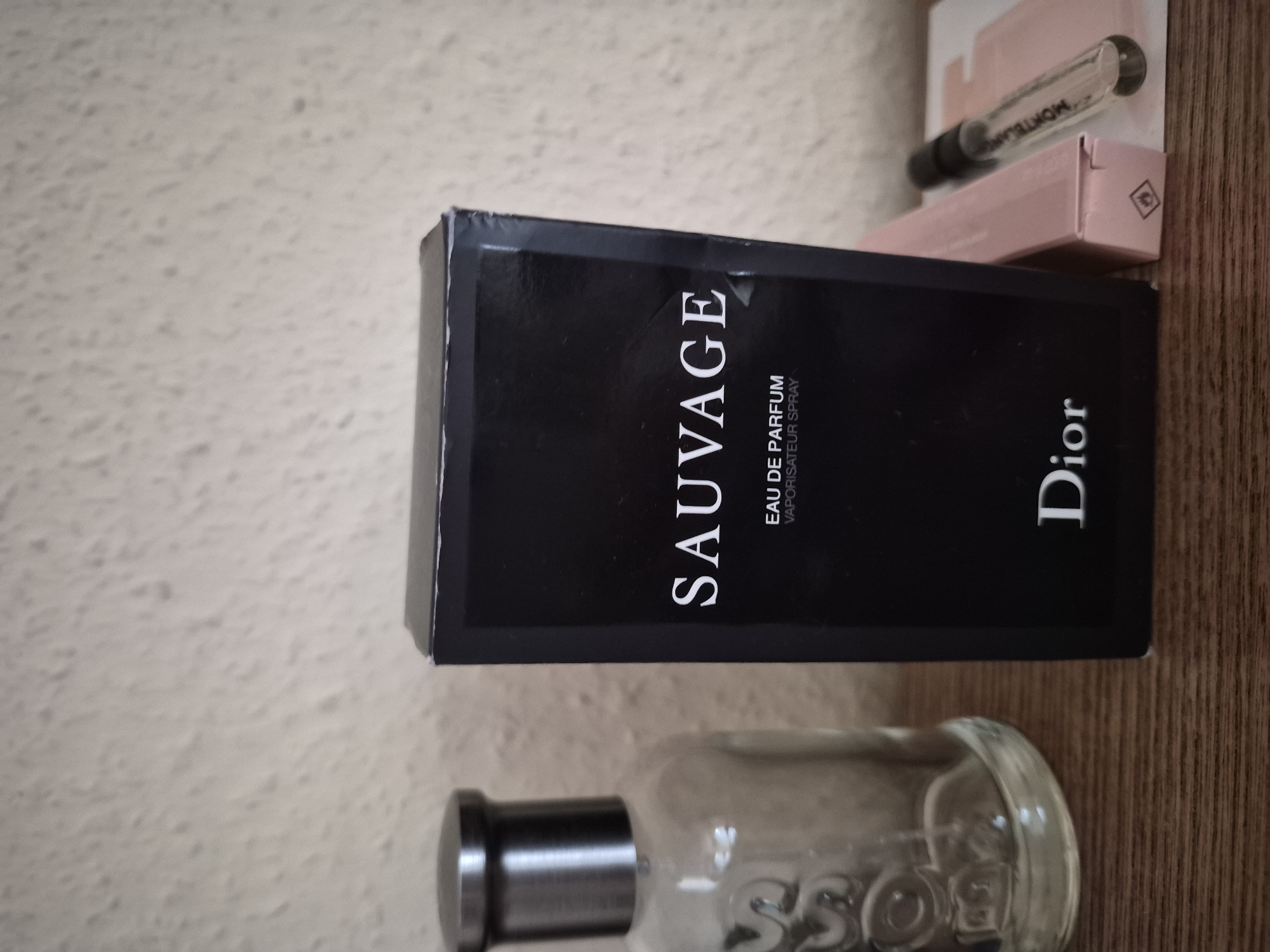 An overhead close-up shot shows a black box of Dior Sauvage Eau de Parfum next to a clear glass bottle of Boss bottled fragrance. The box is upright and positioned in the center of the frame. The glass bottle is to the left of the box and appears to be half empty. To the right of the box are two small, light pink boxes and a clear glass vial with a black cap. The vial contains a clear liquid and has some lettering on it. The objects are placed on a wooden surface. The background is a textured, off-white wall.