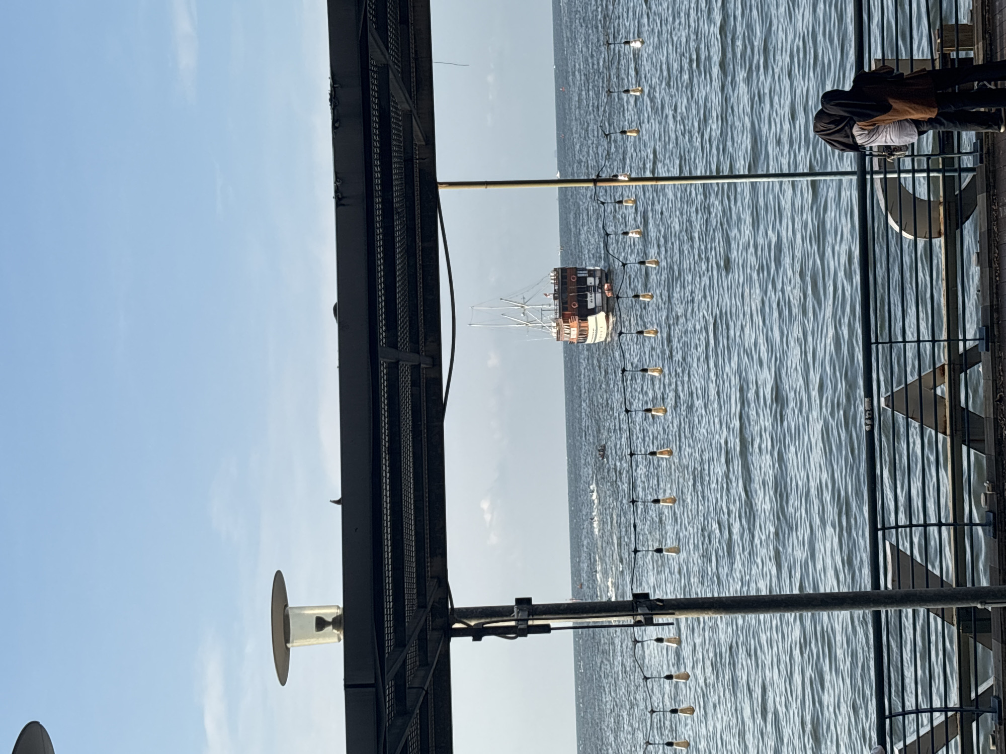 The image depicts a view from a pier or elevated platform in North Jakarta, Indonesia, overlooking the sea. A pirate ship is visible in the distance, situated in the middle of the water. A string of lights hangs along the side of the pier. Two people are standing near a metal railing on the pier. The sky is clear with a few clouds. Structural metalwork is on the left side of the pier.