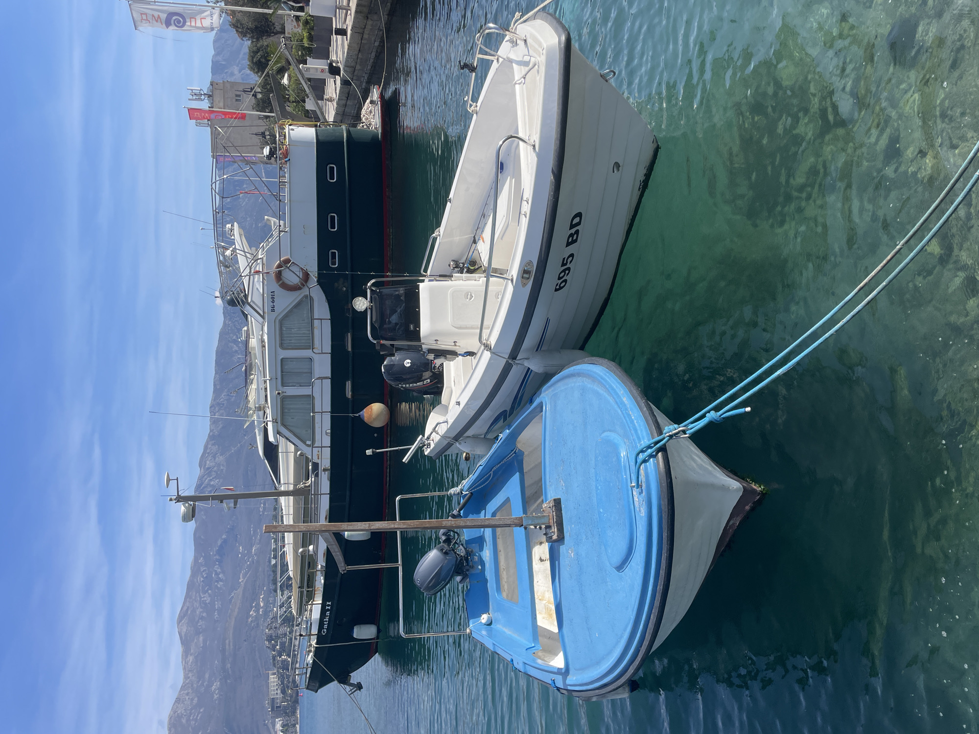 The image depicts a marina scene in Budva, Montenegro. Three distinct boats are moored in clear, greenish-blue water under a blue sky with scattered clouds, with a mountainous and urban coastline in the background.
In the foreground, a blue-decked and white-hulled fiberglass boat is positioned on the left, equipped with a dark outboard motor and a central pole structure. It is secured by a blue rope from its bow extending into the water. To its right, a white fiberglass powerboat displays "695 BD" on its bow, features a black outboard motor, stainless steel railings, and two white fenders. This boat is moored to a stone dock on the right side of the frame and also connected by rope to the blue boat. The water beneath these two boats is transparent, revealing the rocky seabed.
In the midground, a larger dark green and white cabin cruiser, identified as "Gatka II" with registration "BG-601A," is moored. This vessel features multiple cabin windows, a white upper deck with railings, and a mast equipped with radar and communication antennas. Another large white yacht is partially visible behind "Gatka II."
The background reveals a coastline featuring multiple buildings, including what appears to be a multi-story complex, and a tall white tower with red antenna installations. Two flags are visible near the tower, displaying "LUČKA UPRAVA" (Port Authority) and "LUČKA KAPETANIJA" (Port Captaincy) in Montenegrin. Lush green vegetation dots the land, which slopes up to a range of large, barren mountains under the distant sky. No individuals are visible in the scene.