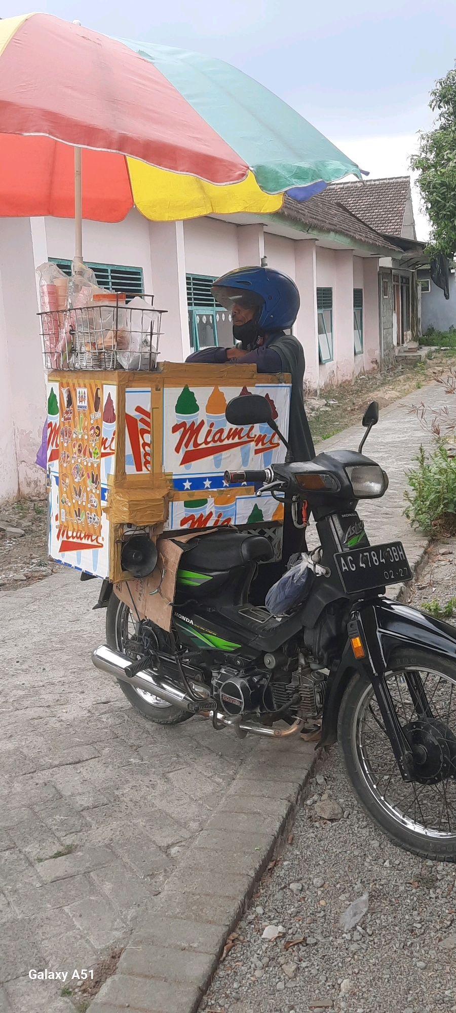 A person wearing a blue helmet stands behind an ice cream cart attached to a motorcycle. The ice cream cart is decorated with images of ice cream cones and the word "Miami". The motorcycle is black and has a license plate that reads "AG 4784 JBH". The scene appears to be in a residential area in Tangkil, Indonesia, with a house visible in the background. The ground is paved with interlocking bricks.