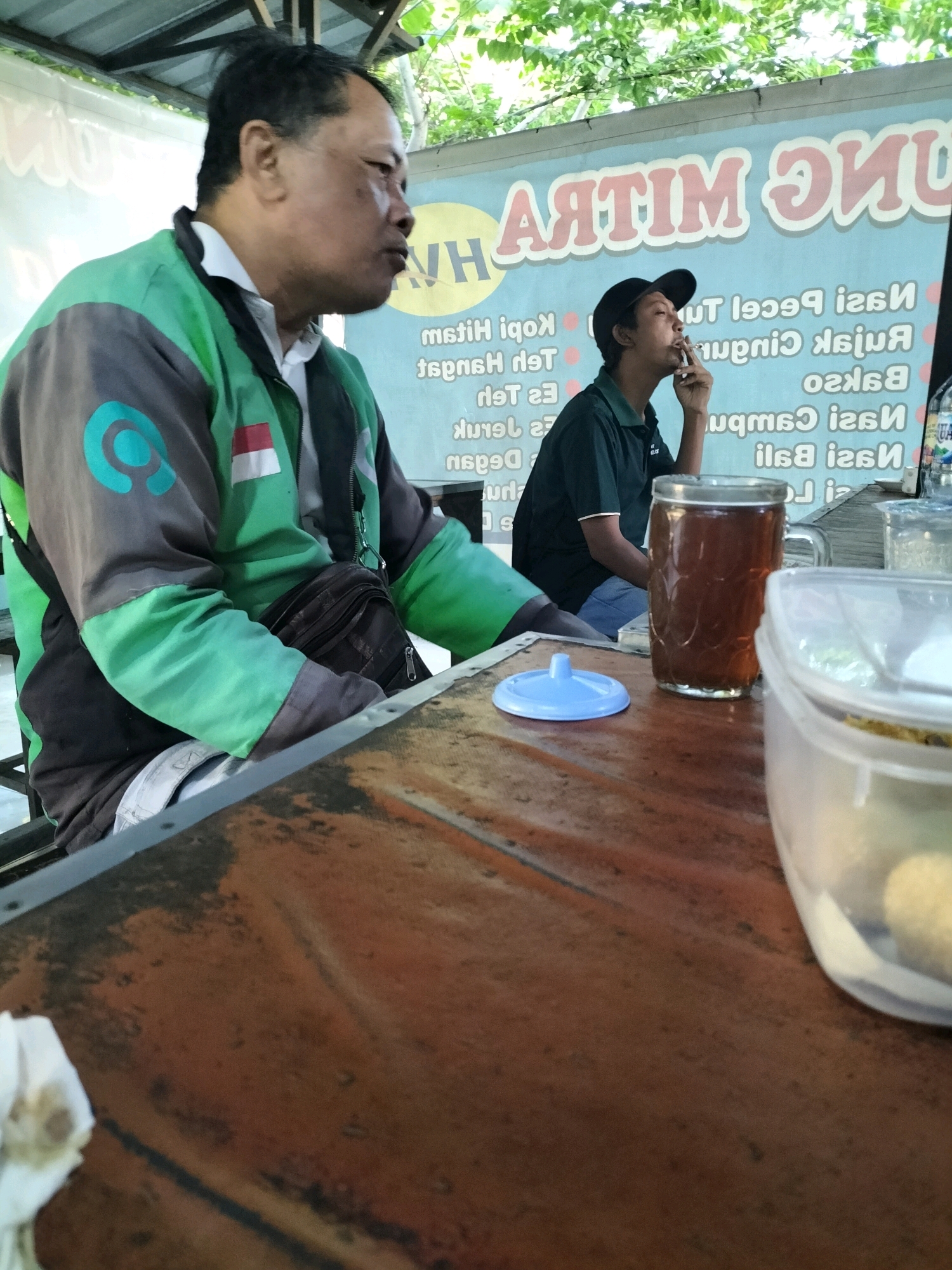 The image shows two men at an outdoor food stall in Pare, Indonesia. On the left, an overweight man in a green and gray jacket with a Gojek logo is seated. He is looking to his right, with his mouth open as if speaking or eating. On the right, a thinner man wearing a black cap and a dark green polo shirt is smoking a cigarette and looking upwards. He is seated behind a table with a glass of iced tea and a plastic container. The table is made of worn wood with a rusty metal edge. In the background, a blue sign with white lettering is visible, along with some greenery from trees. The overall environment suggests a casual dining setting.