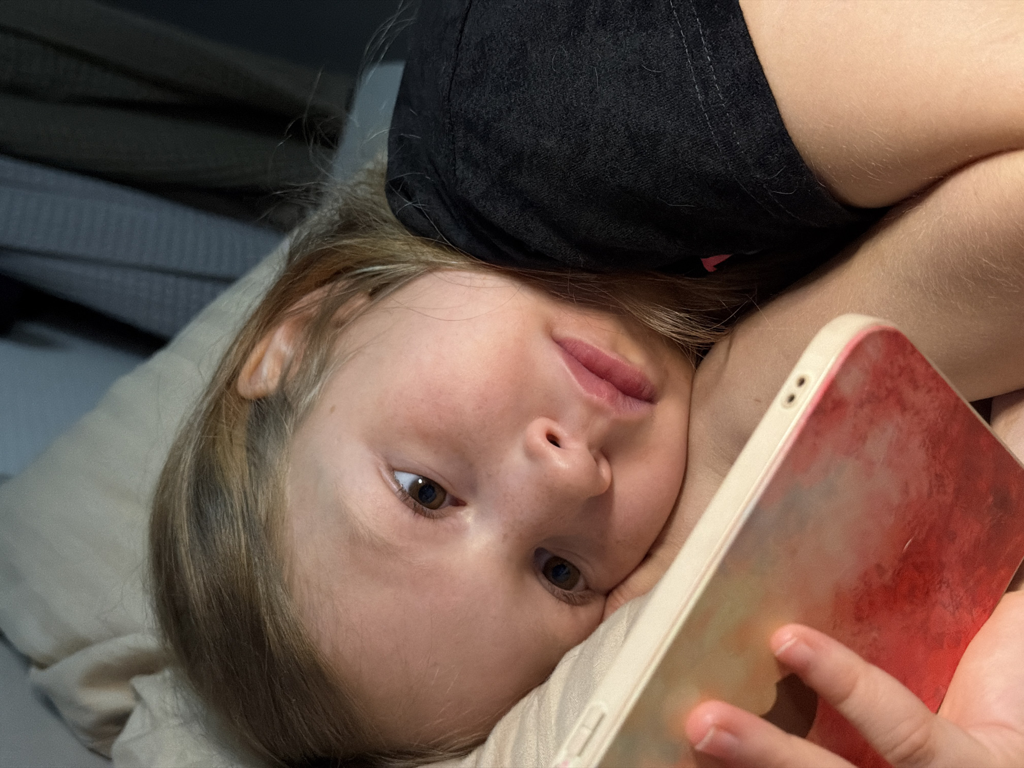 A young fair-skinned child with brown hair and eyes is lying on a light-colored pillow. They are wearing a dark t-shirt and holding a smartphone with a red and gold-colored case. The background is dark, possibly a wall or headboard. The image suggests the child is indoors and possibly in bed. There is no information in the image to confirm the location in the city of Dubovaya, Russia.