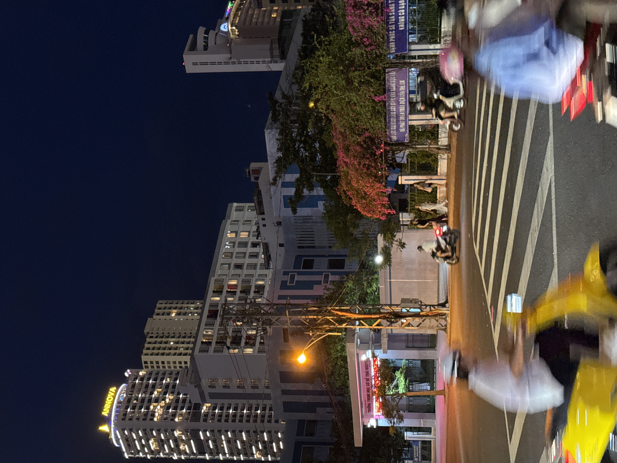 In Nha Trang, Vietnam, at night, a busy street scene unfolds in front of a school entrance. The "ANNOVA" sign glows atop a tall, modern building to the left, which is illuminated with many lights. To the right, other buildings are visible, including a tall, white structure with horizontal lines, and further back, a brighter building with more lights. The school entrance features a white archway with red neon lettering, partially obscured by trees. A street light casts an orange glow on the scene. Several individuals are on motorbikes, their movement captured as blurs due to motion. One person in a white shirt is on a yellow motorbike in the foreground, crossing a zebra crossing. Another person in a blue shirt on a motorbike is also blurred in the foreground. In the background, more motorbikes and pedestrians are visible near a fence adorned with pink flowers. Banners with Vietnamese text are hung along the fence. The sky is a deep, dark blue.