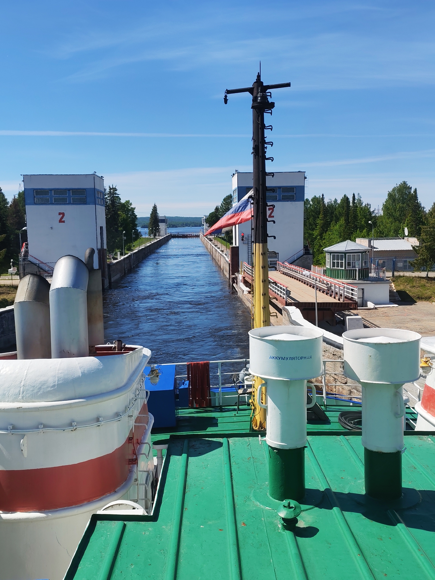 This media file depicts a ship navigating a canal lock on a clear, sunny day. The view is from the deck of the vessel, looking forward into the waterway.
In the immediate foreground, the ship's green deck is visible, featuring various maritime equipment. On the left, a portion of a white and red striped life raft or boat is seen, alongside two large, white and silver colored vents or smokestacks. On the right, two white and green cylindrical structures, likely ventilation shafts, rise from the deck; one clearly displays the Cyrillic text "АККУМУЛЯТОРНАЯ," meaning "BATTERY ROOM" or "ACCUMULATOR." A black mast, from which a Russian flag is flying, stands centrally on the deck.
The ship is positioned within a narrow canal with deep blue water, indicating it is either entering or exiting a lock. Flanking the canal are tall, white lock towers with blue trim, each prominently marked with a large red number "2." On the right bank, a small building with a green octagonal roof and a wooden gangway leading to it are visible. Dense green forests line both sides of the canal, extending into the distance.
In the background, beyond the lock, a wide body of water, possibly a lake or a larger river, can be seen under a bright blue sky dotted with faint, wispy clouds. The scene conveys a sense of peaceful transit through a regulated waterway. The bright sunlight creates strong shadows and highlights the clear weather conditions.