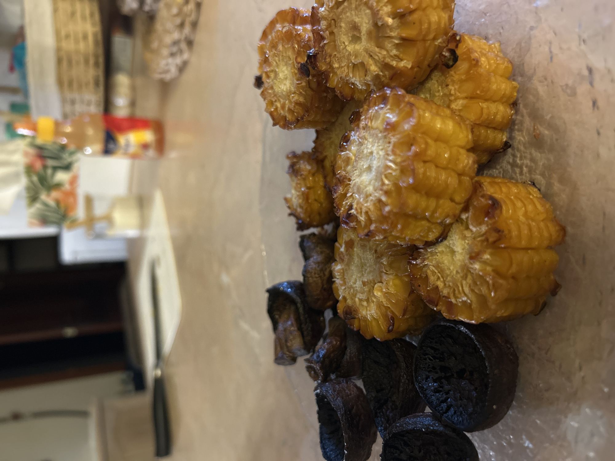 The image shows a plate of grilled corn on the cob cut into segments and what appear to be grilled mushroom slices. Some of the mushroom slices appear to be charred. The plate rests on a table with other blurred objects in the background, including what may be bottles and other kitchen items. The location is assumed to be Chaikovsky, Russia, but there is no information in the image itself to verify this. There are no people visible in the image.