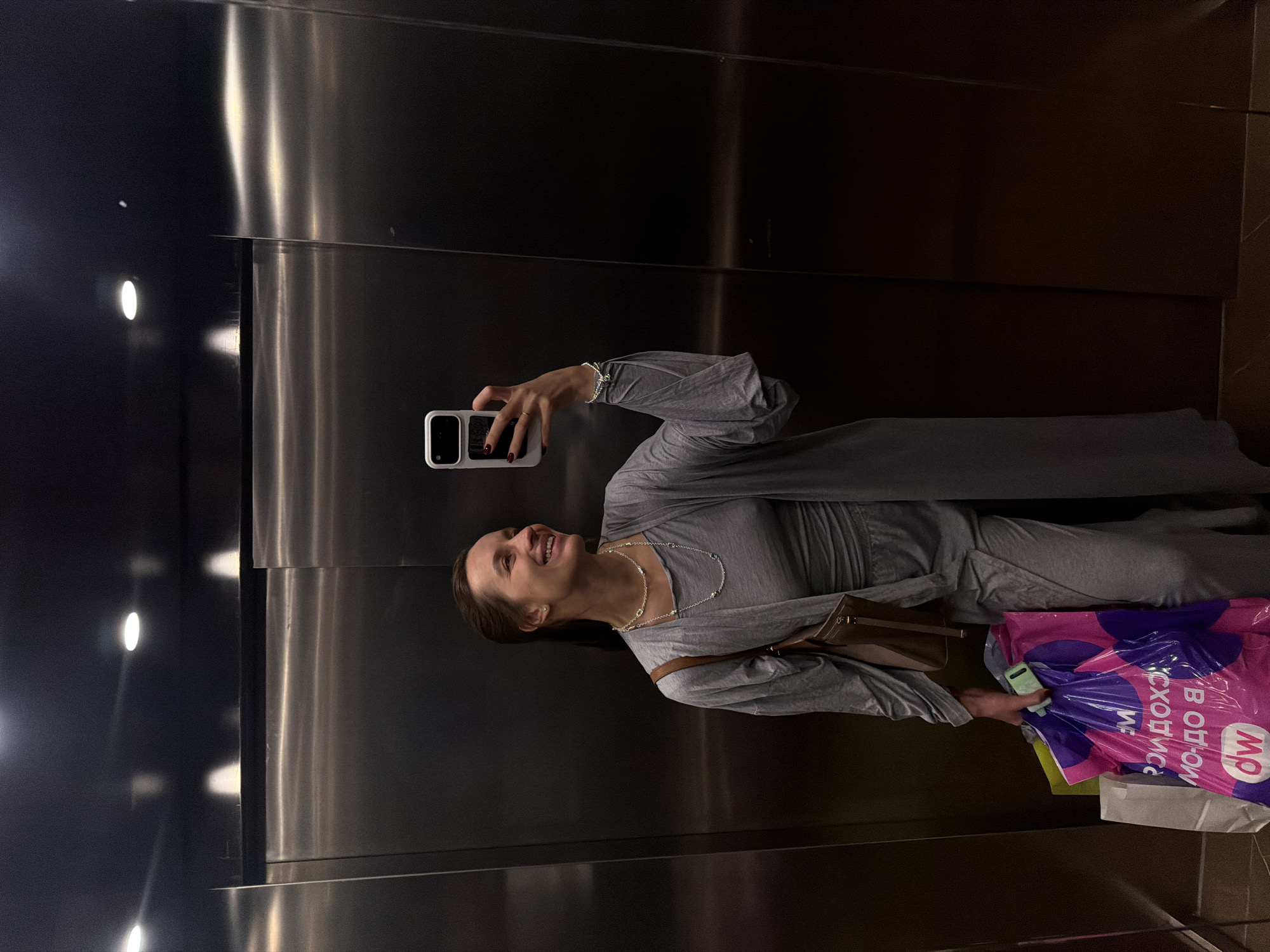A woman is standing in an elevator, holding a smartphone in her left hand as if taking a selfie. She is wearing a gray outfit, a silver necklace, and a bracelet on her left wrist. In her right hand, she holds shopping bags, including one with the recognizable logo of the "Московский" department store, indicating a location in Moscow, Russia. The interior of the elevator is sleek and modern, with metallic surfaces and recessed lighting.