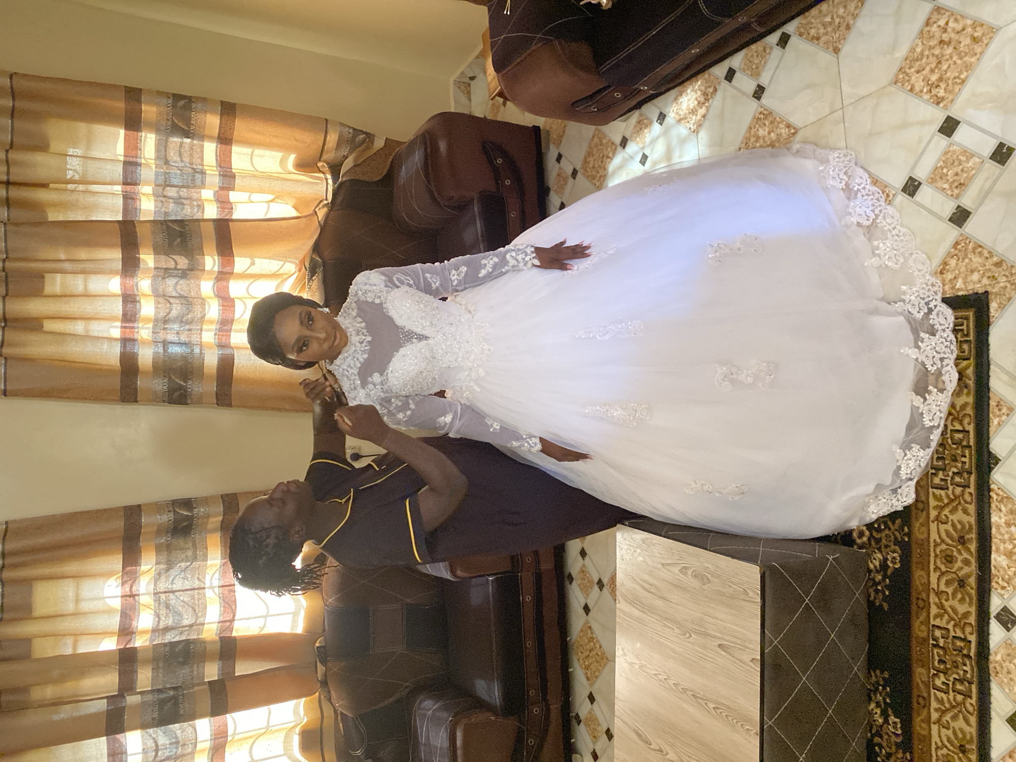 A woman in a white wedding gown is having her makeup applied by another woman. The woman applying the makeup is wearing a dark-colored uniform with yellow trim. They are positioned in front of a window with patterned curtains. The setting appears to be indoors, in a space with brown leather furniture and patterned flooring. It is likely Bukuru based on the prompt.
