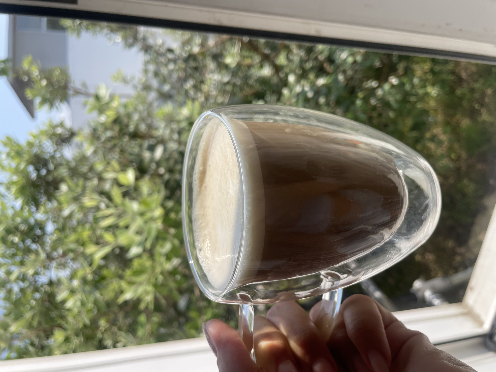 A close-up, eye-level shot shows a hand holding a double-walled glass mug filled with coffee, with a creamy froth on top. The mug is positioned in the foreground, with a blurred background of green foliage and a white building, suggesting an outdoor or window-facing setting. The lighting is bright and natural, casting subtle shadows. The image appears to be captured in Baranovka, Russia, based on the provided context.