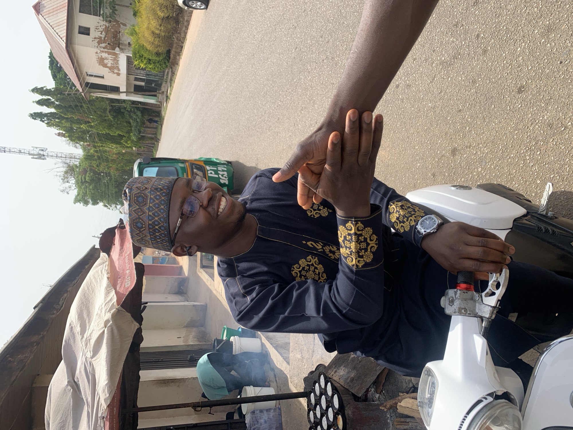 A man wearing a blue and gold patterned top and a matching kufi cap sits on a white scooter on a city street in Jos, Nigeria. He is smiling and shaking hands with someone out of frame. A watch is visible on the man's left wrist. In the background, a green "keke" tricycle taxi is parked, and a dilapidated building is seen further down the street. To the left of the man, there is a cooking operation with smoke rising, suggesting food is being prepared outdoors. Buckets and cooking equipment are visible next to the cooking area.
