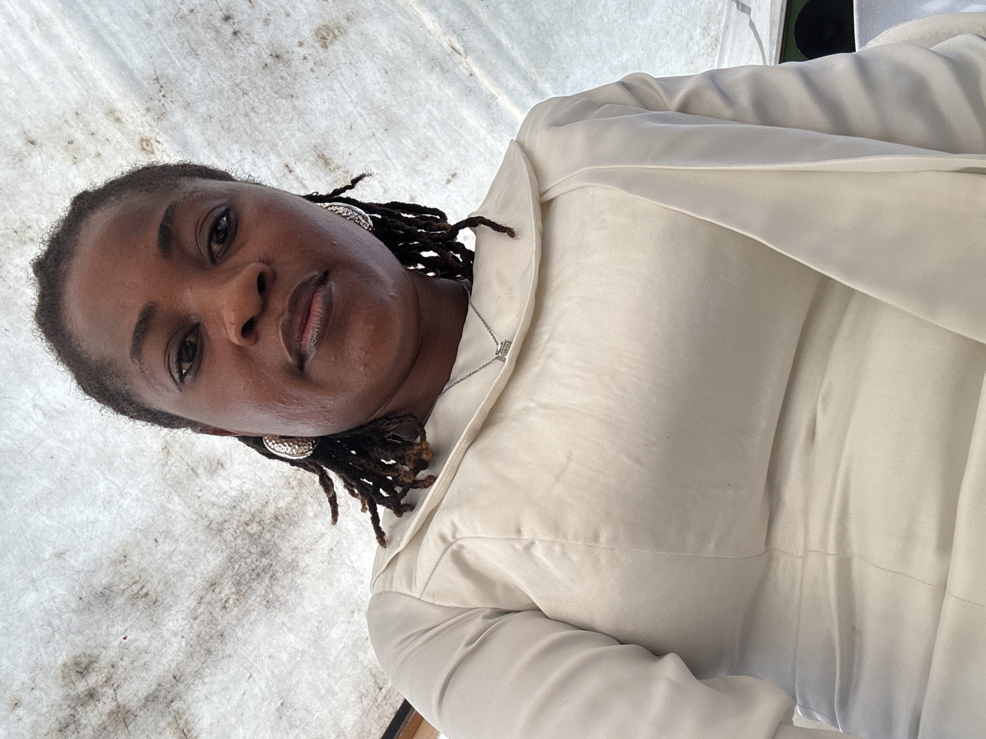 A medium-shot, low-angle, eye-level selfie shows a woman with dark skin and dreadlocks, wearing a cream-colored dress with a wide collar, and silver hoop earrings. She is looking directly at the camera with a neutral expression. The background is a white, textured surface that appears to be a tarp or tent ceiling, possibly in an outdoor setting in Badagry, Nigeria. The lighting is bright, suggesting daylight.