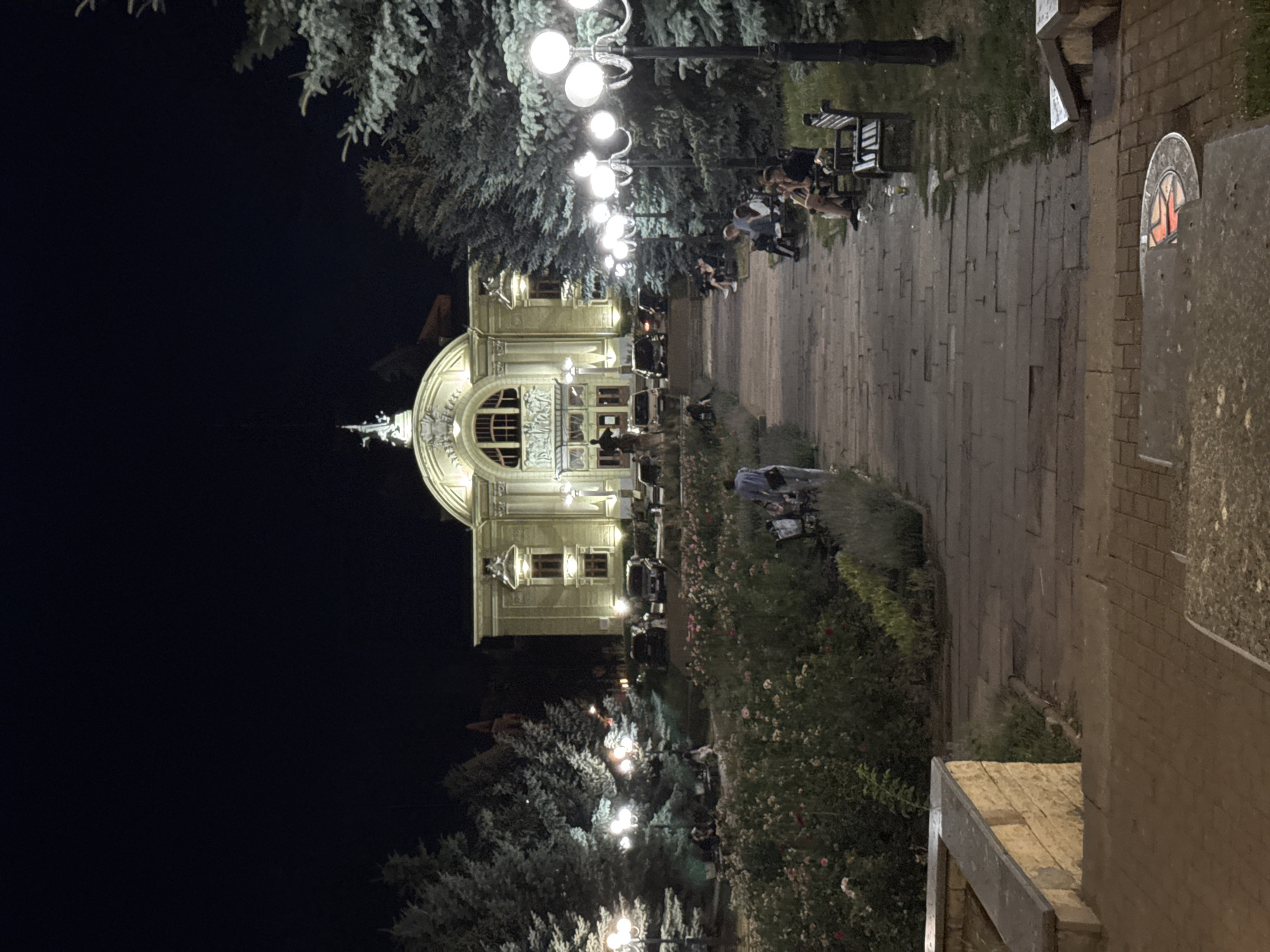 This is a nighttime photograph of the Chernivtsi Opera and Ballet Theatre in Chernivtsi, Ukraine. The imposing facade of the theatre is illuminated by spotlights and streetlights, casting a warm glow on the ornate architecture. A pathway lined with benches and lampposts leads towards the entrance. Several people are seated on the benches or walking along the path. In the foreground, a flower bed with blooming roses adds a touch of color to the scene. The overall atmosphere is one of a peaceful evening in a European city square.