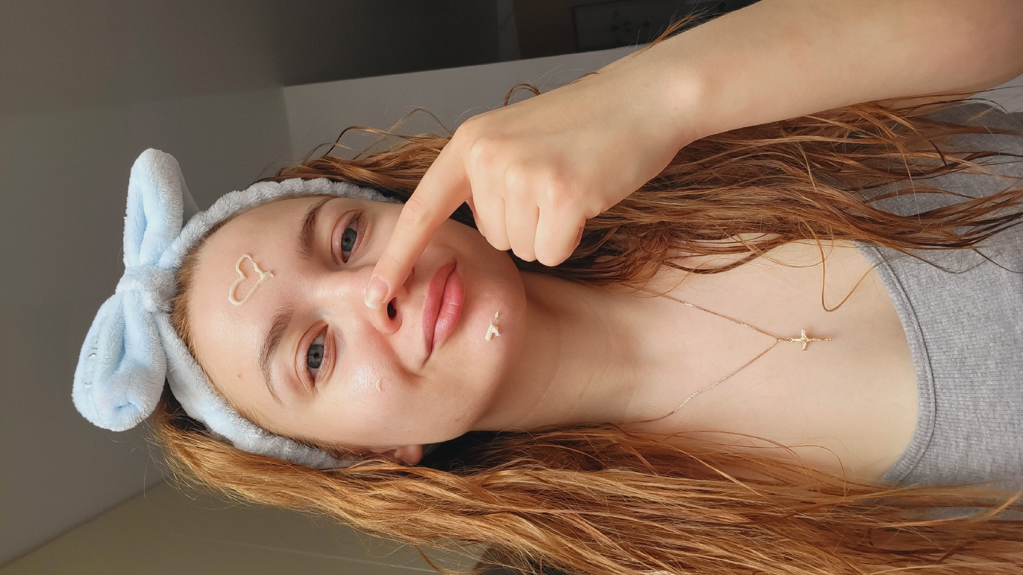 A young woman with long, wavy auburn hair is facing the camera. She is wearing a light blue headband with bunny ears. There are dabs of white cream on her forehead, nose, and chin. She is touching her nose with her index finger. She is wearing a light grey shirt and a delicate gold necklace with a cross pendant. The background is blurred and appears to be a room interior with neutral-colored walls. There is no information available in the image to confirm the location as Chaikovsky, Russia.