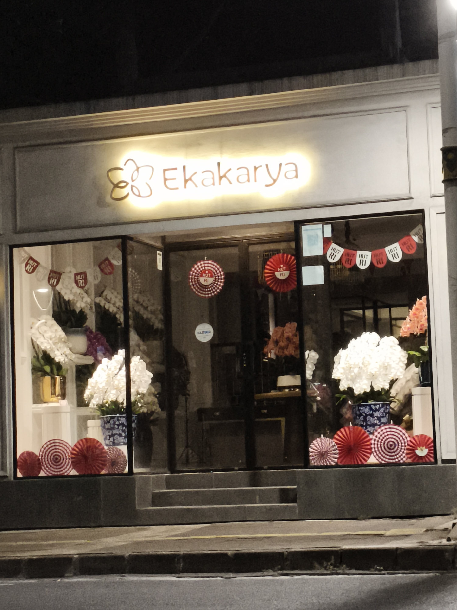 This is a night time, eye-level, outdoor, medium shot of a storefront with a brightly lit sign that reads "Ekakarya" with a floral design. The storefront has large glass windows displaying floral arrangements and decorative paper fans in red and white patterns. The word "CLOSED" is visible on a sign inside the window. The street in front of the store is visible, with a sidewalk and stone steps leading up to the entrance. The overall atmosphere is festive and inviting, suggesting a shop that sells flowers or gifts. The location of the store is in the city of South Jakarta, Indonesia.