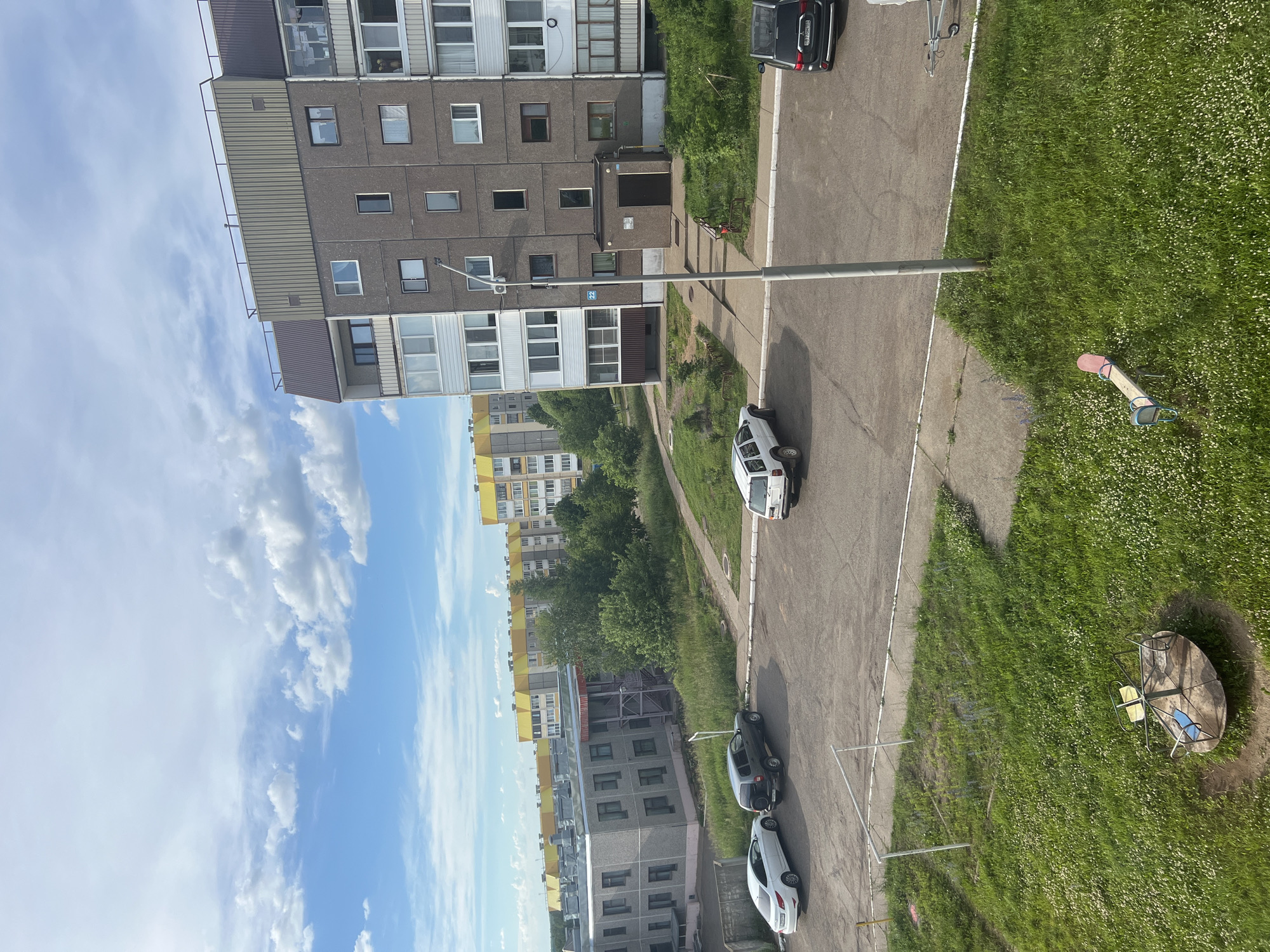 The image depicts a view of a residential area in Markovsky, Russia, under a partly cloudy sky. In the foreground, a grassy area with a merry-go-round and a seesaw is visible, with a paved pathway and parking area adjacent to it. Several parked cars, including a white sedan, a dark SUV, and a white SUV, are present in the parking lot.
Further into the scene, a multi-story apartment building dominates the right side of the frame. This building has a textured gray facade, with numerous windows and balconies. A street lamp stands in front of the building. Across from the apartment building, a lower, gray building with multiple windows is visible, likely a school or another community building. In the background, several taller apartment buildings with yellow and gray facades stretch across the horizon. The environment is characterized by urban residential structures interspersed with green spaces and trees.