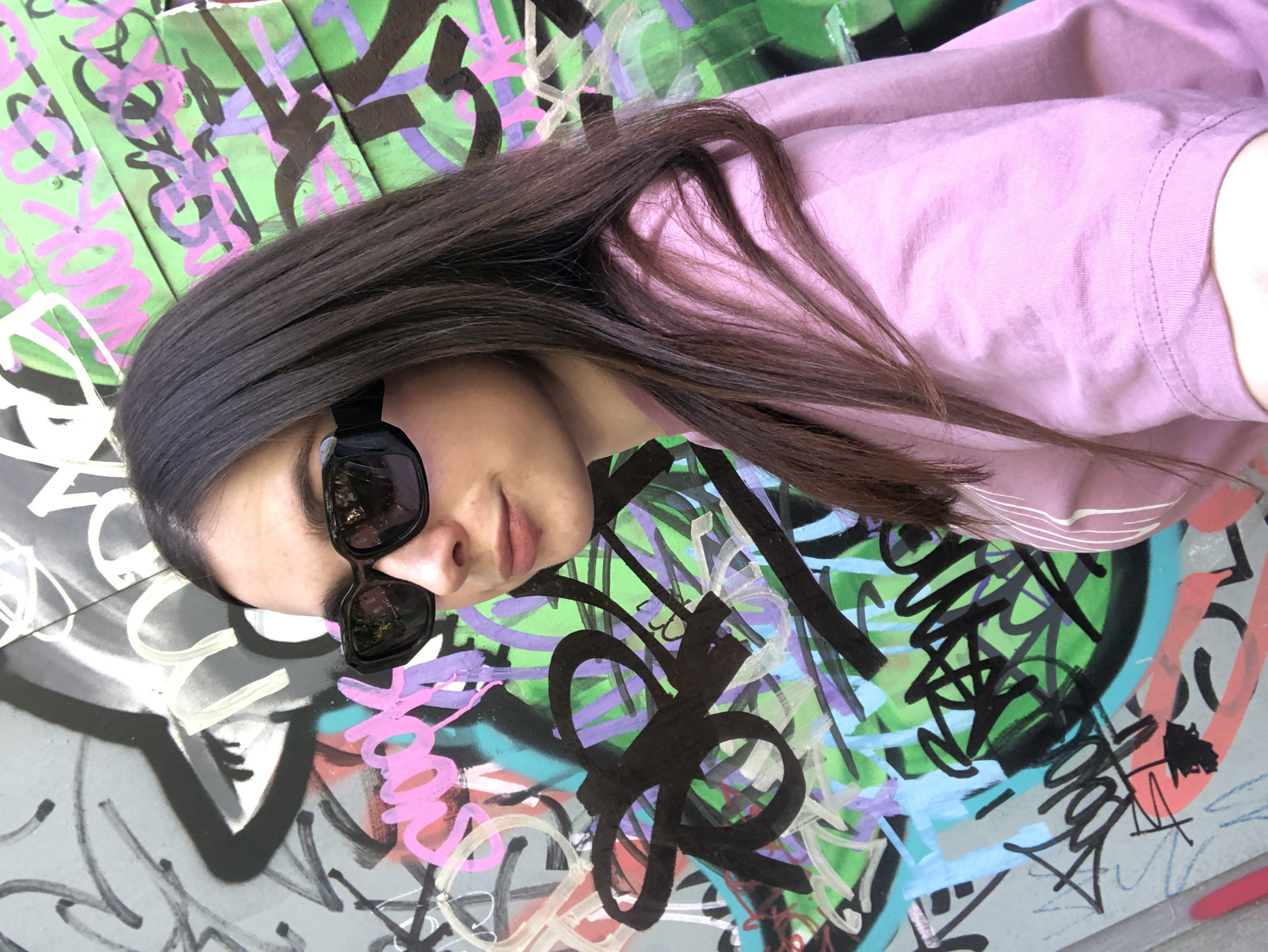 A young woman with long, dark hair and sunglasses is seen in a close-up shot against a backdrop of graffiti. She is wearing a light pink shirt. The graffiti behind her includes a mix of colors like green, purple, black, and light blue, with various abstract patterns and shapes. The image is rotated, with the woman appearing on her side. Based on additional information, this scene is located in the city of Chaikovsky, Russia.