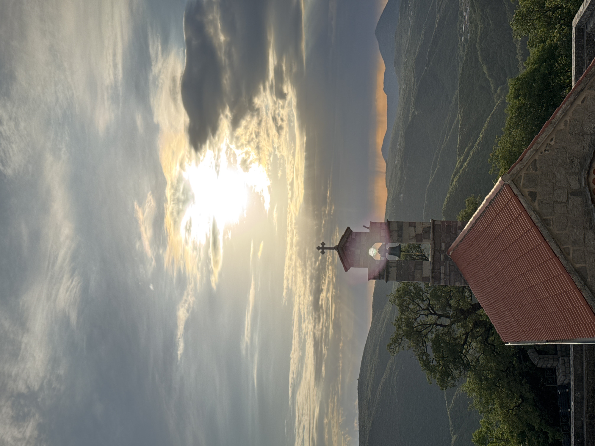The image depicts a landscape at sunset featuring a stone bell tower with a red-tiled roof, situated in the location of Bratešići, Montenegro. The bell tower has a cross atop its roof. The sun shines brightly through clouds and is adjacent to the bell tower. Mountainous terrain appears in the background. Trees and foliage surround the bell tower. The sky exhibits both blue and cloudy conditions.