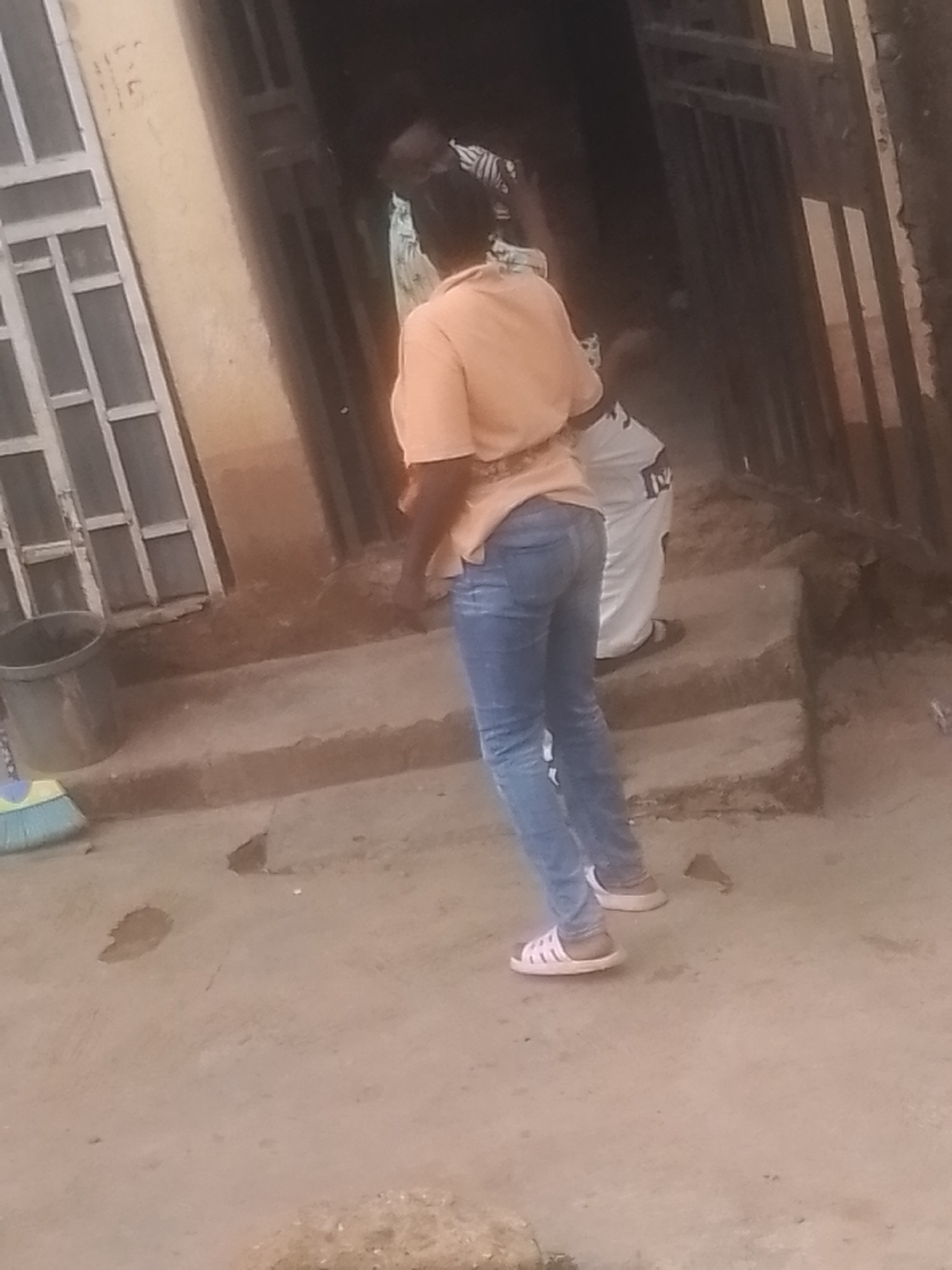 The image shows two people standing on a concrete surface in front of a building with a dark entrance and a window on the left. A person in the foreground is wearing a light orange t-shirt and blue jeans, with white sandals. Their back is to the camera, and they appear to be looking towards the building. Behind them, another person is partially visible, wearing a white garment with black markings. A gray bucket and a blue and yellow broom are on the left side of the image, near the building's entrance. The ground appears to be dusty and there are some small puddles or stains on it. The lighting suggests it might be daytime, but the overall scene is somewhat dimly lit, possibly due to the shadows cast by the building or overcast weather. The location cues, such as the building's architecture and the dusty ground, are consistent with a setting in Gwong, Nigeria. There is no visible text in the image. The activity appears to be mundane, with the individuals either standing around or possibly about to engage in some task, given the presence of the broom and bucket.