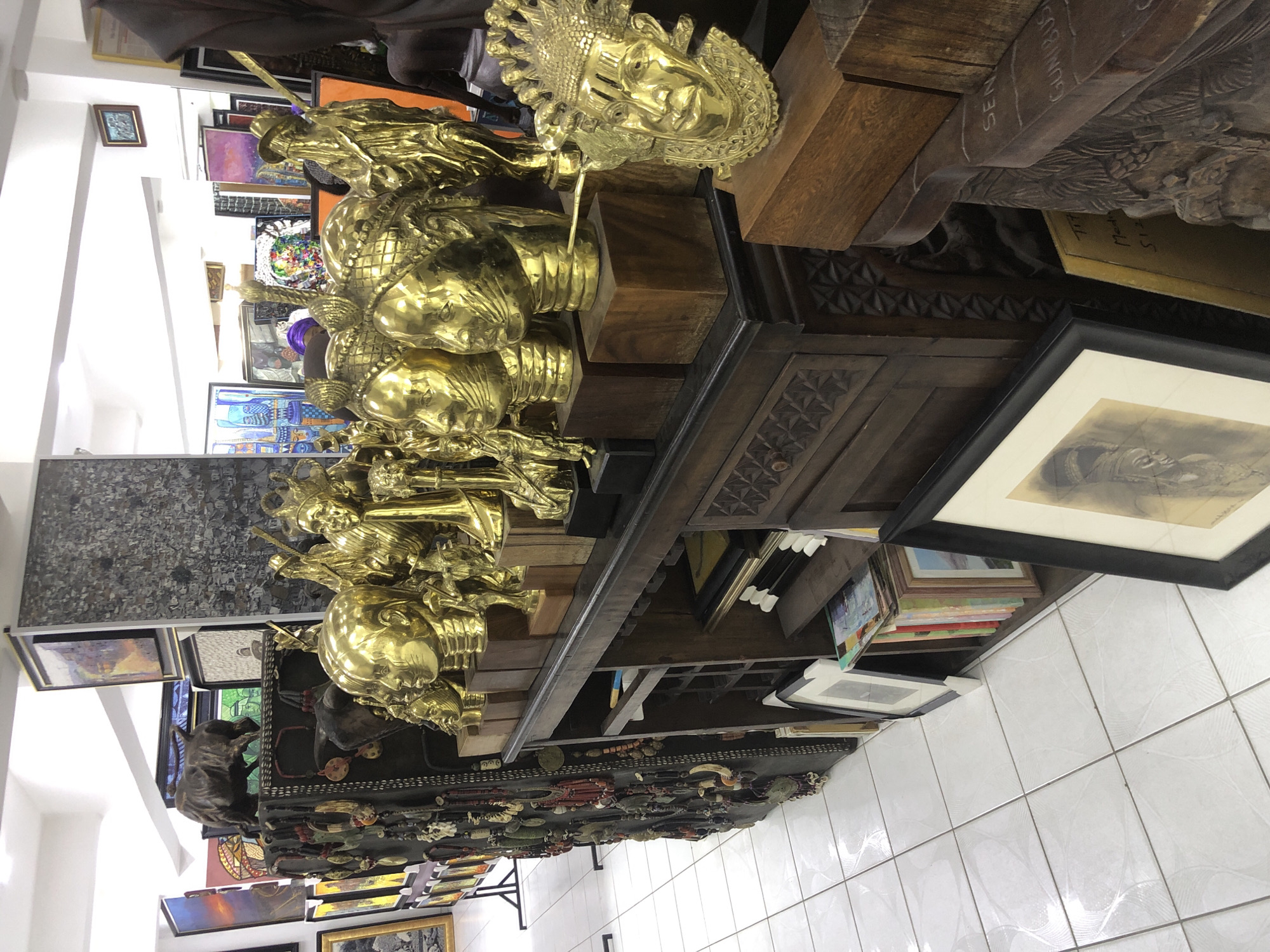 The image depicts a retail display of artisanal goods, likely in a shop or market stall, in Alaguntan, Nigeria. The foreground is dominated by a collection of polished gold-toned busts, primarily of human heads, displayed on a dark wooden cabinet. These busts feature intricate details, with some wearing elaborate headpieces or jewelry. To the left of the busts, a dark, textured surface is adorned with numerous necklaces and decorative pendants, suggesting a display of traditional adornments.
Behind the gold busts, a dark wooden cabinet with carved details holds stacks of framed artwork and books on its shelves. One framed artwork in the lower right corner features a black and white sketch of a person's head. The overall environment appears to be an indoor setting with white tiled flooring. The background reveals more artwork, including paintings with vibrant colors and abstract designs, suggesting a diverse range of crafts and artistry. The lighting is bright, illuminating the polished surfaces of the gold artifacts.