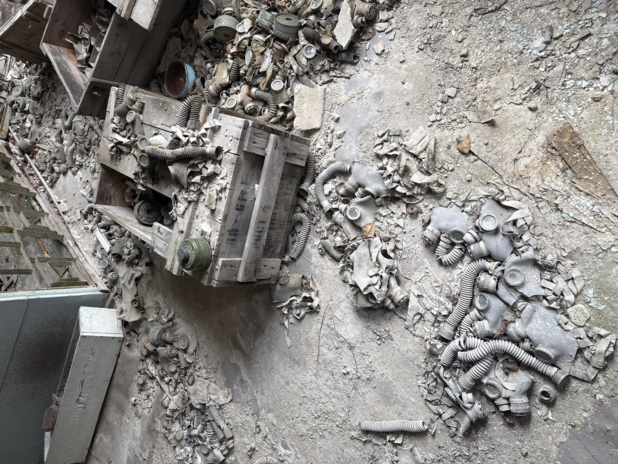 The image depicts the interior of a building in Prypiat, Ukraine, filled with discarded gas masks. Multiple wooden crates lay overturned, spilling out the masks. The floor is littered with debris and fragments of the masks. The environment is dusty, decayed, and reflects the abandonment of the location. No people are present.