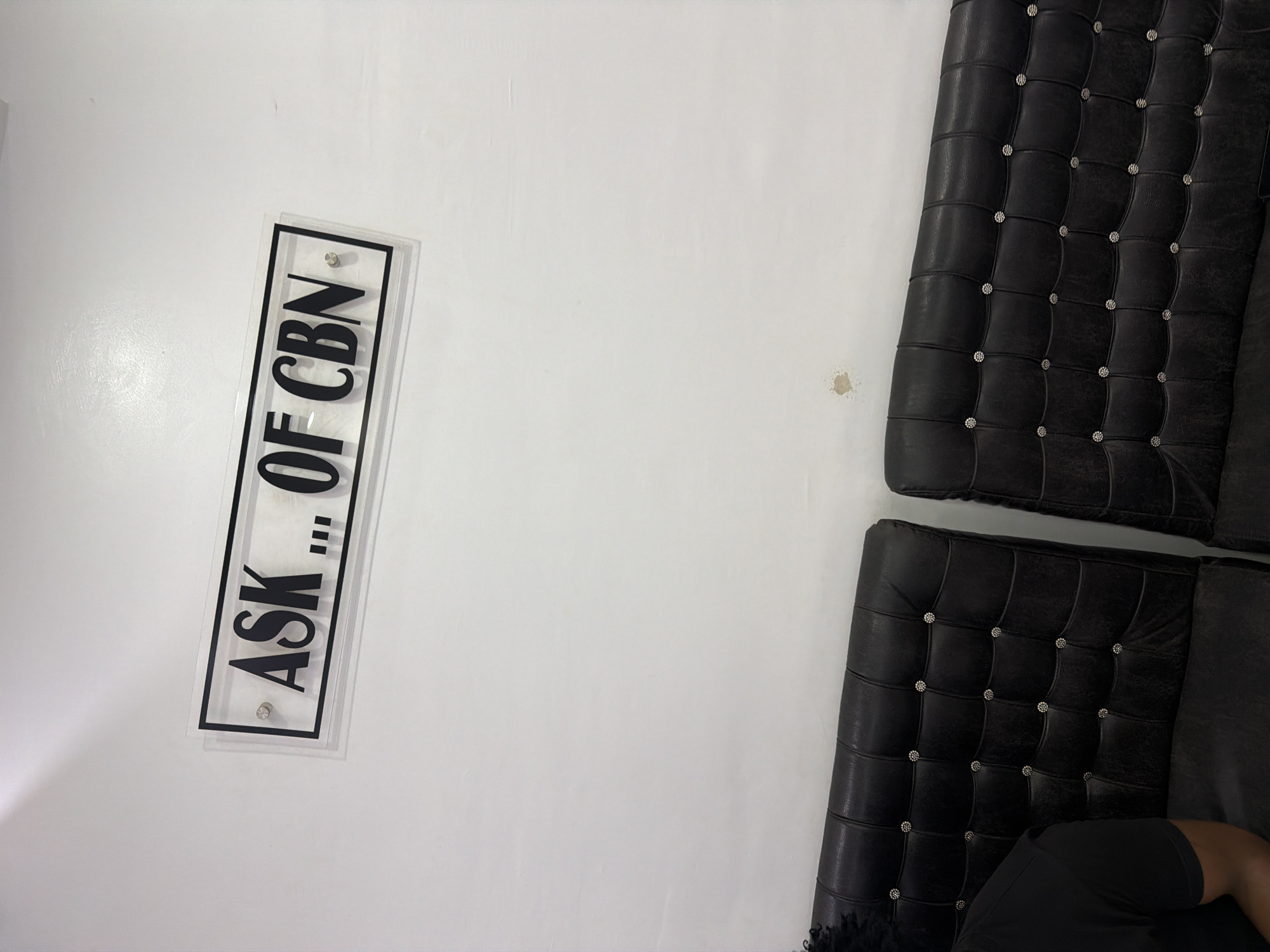 The image shows a close-up, angled view of a white wall with a sign and furniture. The sign is black text on a clear acrylic plate, mounted on the wall. The text reads "ASK,, OF CBN". The sign is oriented vertically. To the right of the sign, two sections of a dark, tufted upholstered couch are visible. The couch appears to be made of leather and has silver button accents. A person's arm and shoulder, wearing a black t-shirt, are visible in the lower right corner, suggesting they are seated on the couch. The lighting suggests an indoor setting, possibly during the day, with a bright light source casting a faint shadow on the wall near the sign. There are a couple of small, discolored marks on the white wall to the left and right of the couch. The overall impression is of a modern, minimalist interior space with a focus on the sign and the furniture.