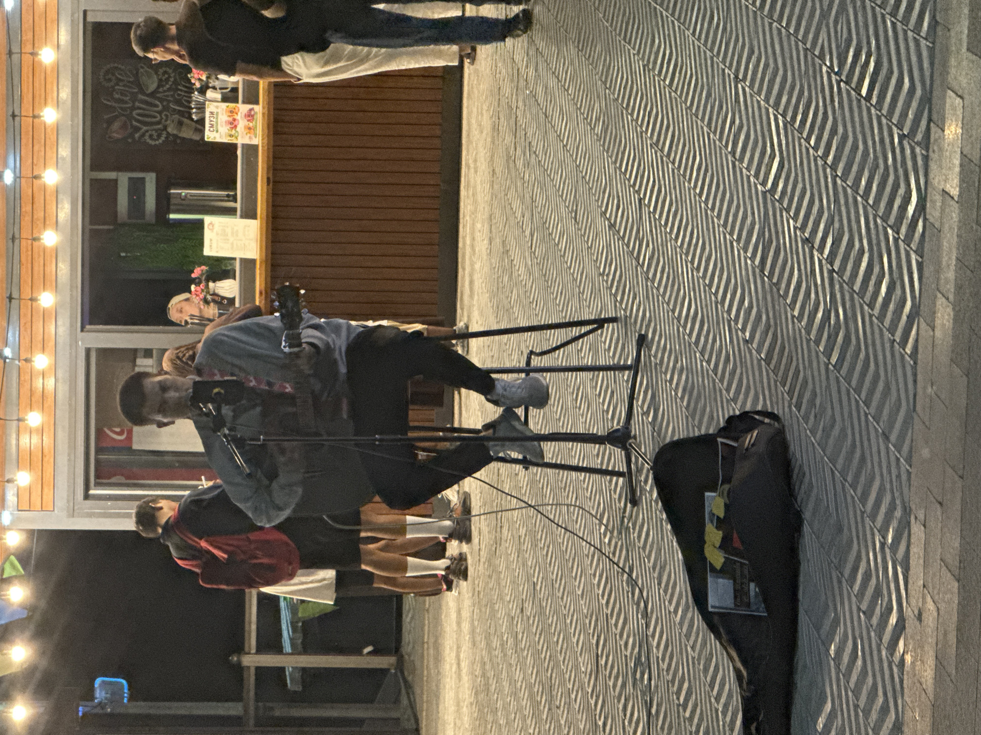 The image depicts an outdoor scene at night, possibly a waterfront or promenade, with a street performer playing a guitar and singing into a microphone. He is seated on a stool in front of a kiosk that appears to be selling food or drinks. Several people are visible in the background, some queuing at the kiosk and others standing around. The ground is paved with a patterned tile. A guitar case lies on the ground in the foreground. The overall atmosphere suggests a casual, public gathering. The image may have been taken in Pavlodar, Kazakhstan, but this cannot be definitively confirmed from the visual information alone.