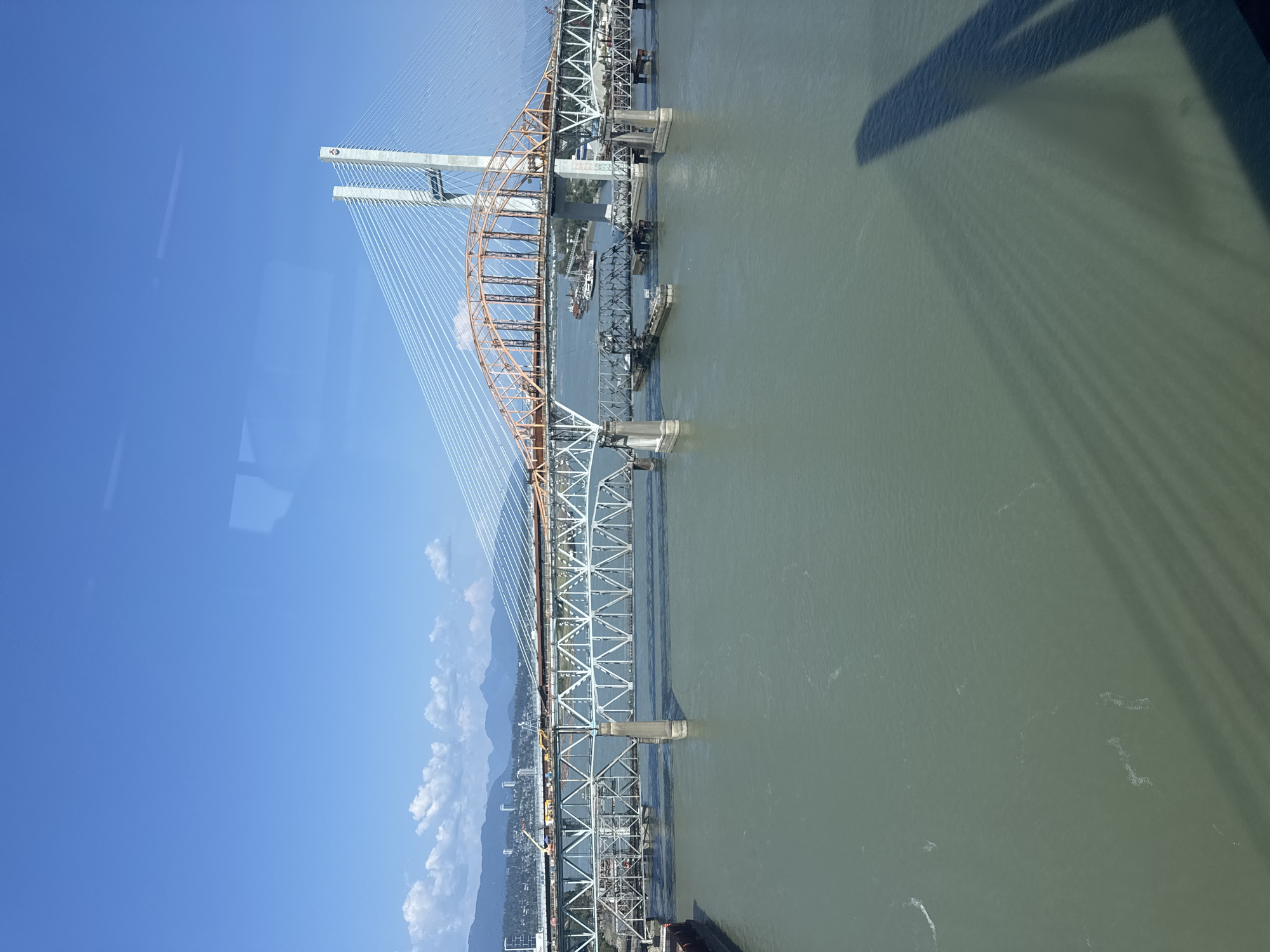 This is a photograph of the Alex Fraser Bridge in New Westminster, Canada, taken on a clear, sunny day. The bridge, a cable-stayed structure, spans a wide, greenish-gray river. Multiple lanes of traffic are visible on the bridge, which is supported by tall white towers and a network of cables. In the background, a distant city skyline with mountains behind it can be seen. The water below the bridge is calm, with some small boats and barges present. A shadow of the bridge or another structure is cast across the water in the foreground. The image appears to have been taken from a slightly elevated position, possibly from another bridge or a tall building.