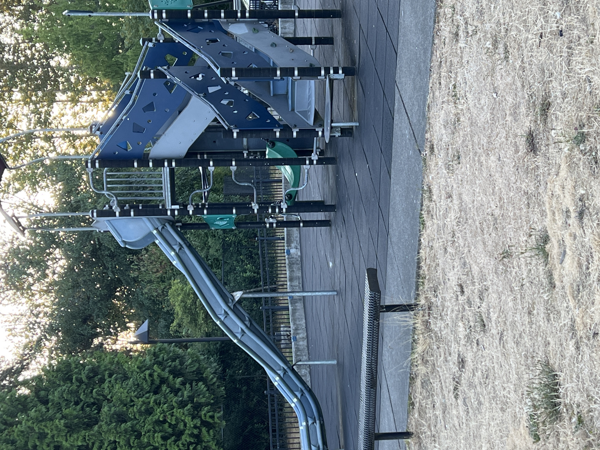 The image captures a playground area on a bright day. In the foreground, a metal bench with a slatted seat is positioned on a dark, rubberized safety surface. Beyond the bench, the safety surface continues, forming a grid pattern. The main subject of the image is a large, multi-level playground structure with slides and climbing elements. The structure features a large, curving blue and gray slide that extends from a higher platform down to the ground level. Other parts of the structure include staircases with blue and gray panels, railings, and a green swing visible in the background. The playground is surrounded by a black metal fence. Behind the fence, a concrete barrier is visible, and further back, dense green trees form a natural backdrop. The sun is shining, casting dappled light through the trees, and the sky is not visible. The overall scene depicts an outdoor recreational space. The location is in Kent, Washington, United States of America.