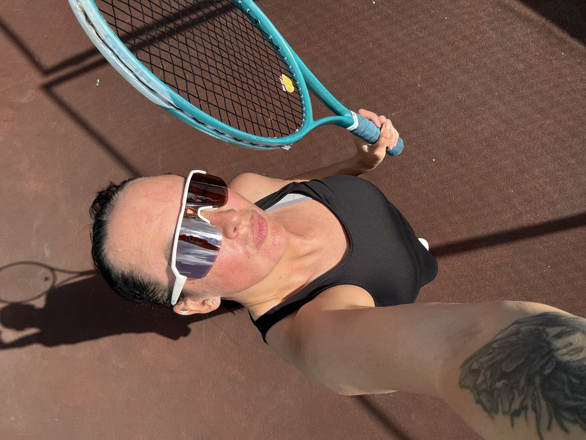 A person is holding a tennis racket overhead, their face turned towards the camera and wearing large white sunglasses. The sunglasses reflect a landscape with mountains. The person is wearing a black tank top. A tattoo of an eagle is visible on their right arm. The scene is outdoors, with a brown surface that appears to be a tennis court. Shadows suggest it is a sunny day. The image is taken from a high angle, looking down at the person. There is no visible text in the image.