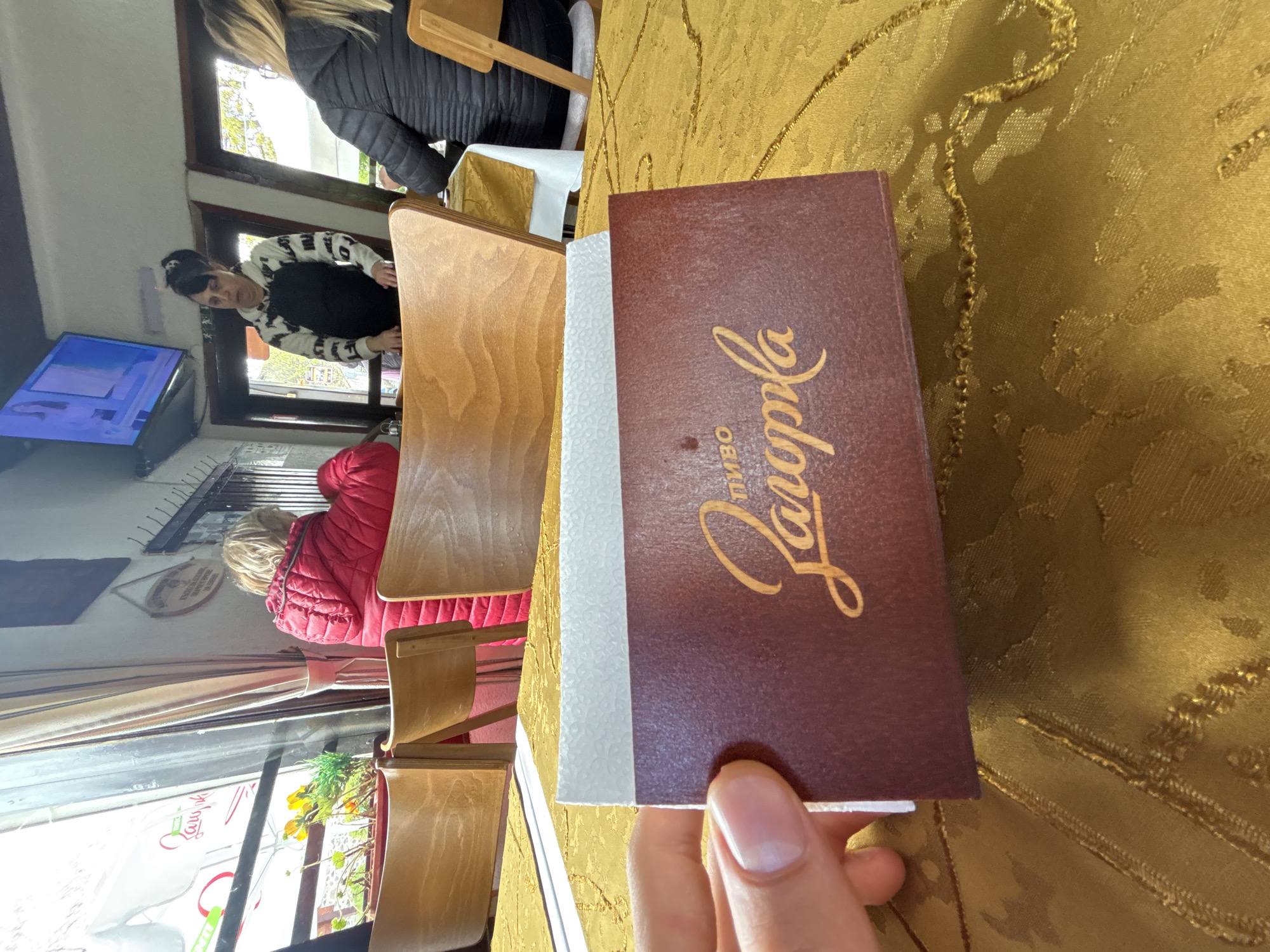 The scene appears to be inside a restaurant or cafe in Zlatograd, Bulgaria. A hand holds a menu card with the Cyrillic word "Јаворла" (Yavorla) written on it. The menu card is brown with a white edge. A golden-colored tablecloth is visible beneath the menu. Seated at wooden tables and chairs are several individuals, including a person in a black jacket, a woman in a patterned sweater, and a person in a red jacket. A television is mounted on the wall and is showing what seems to be a cartoon. A window is also visible.