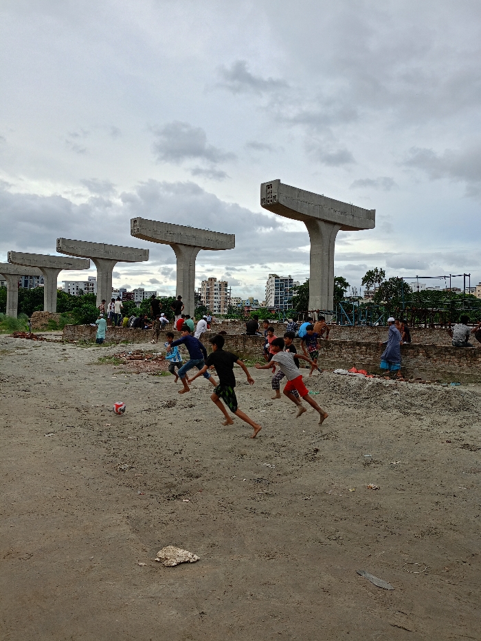 The image depicts an outdoor scene in Sujabad, Bangladesh, characterized by ongoing urban development. In the foreground, five bare-chested or lightly clothed children are playing football on a compacted dirt and sand surface. A red and white football is positioned to the left. Three children are actively running towards the ball; one wears a black shirt, another a grey shirt with red shorts, and a third a blue shirt. In the midground, approximately 15-20 individuals, including adults and children, are gathered, some seated on low brick walls or debris, others standing. Behind them, four large, uncompleted concrete support pillars, typical for an elevated transport system, stand prominently. Residential buildings are visible in the distant background under an overcast sky. Scattered construction debris and remnants of brick structures are present throughout the area.