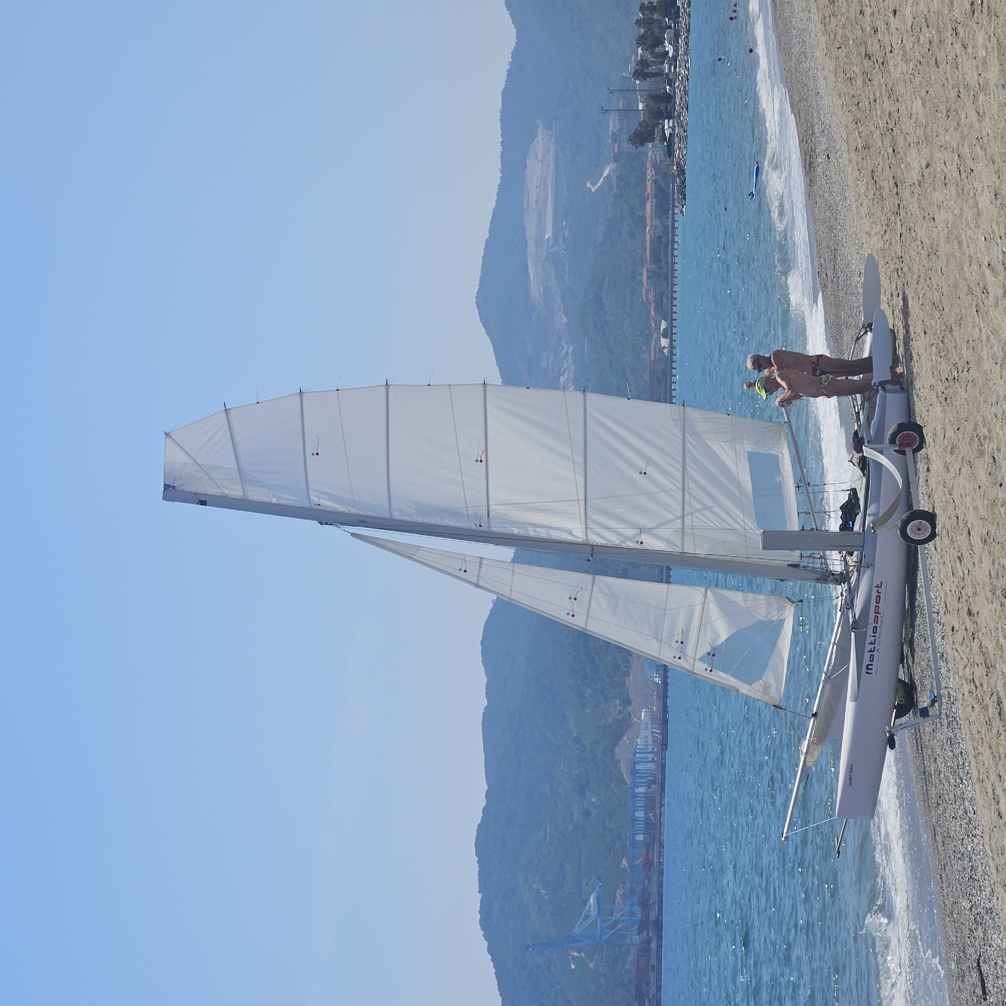In Savona, Italy, a daytime, medium-shot photograph captures a two-hulled sailboat positioned on a pebbly beach adjacent to the Mediterranean Sea. The sailboat, with its white sails raised and secured by rigging, is mounted on a trailer with wheels. Two adult males are interacting with the sailboat. One male, wearing a green cap, stands near the front of the boat, while the other, wearing sunglasses, stands behind him. Both are in swimwear. The sea, a vibrant blue, exhibits small waves breaking near the shore. In the background, a coastline features a port with cranes and industrial structures, and lush, green hills rise behind it. The sky is clear and blue, indicating fair weather.