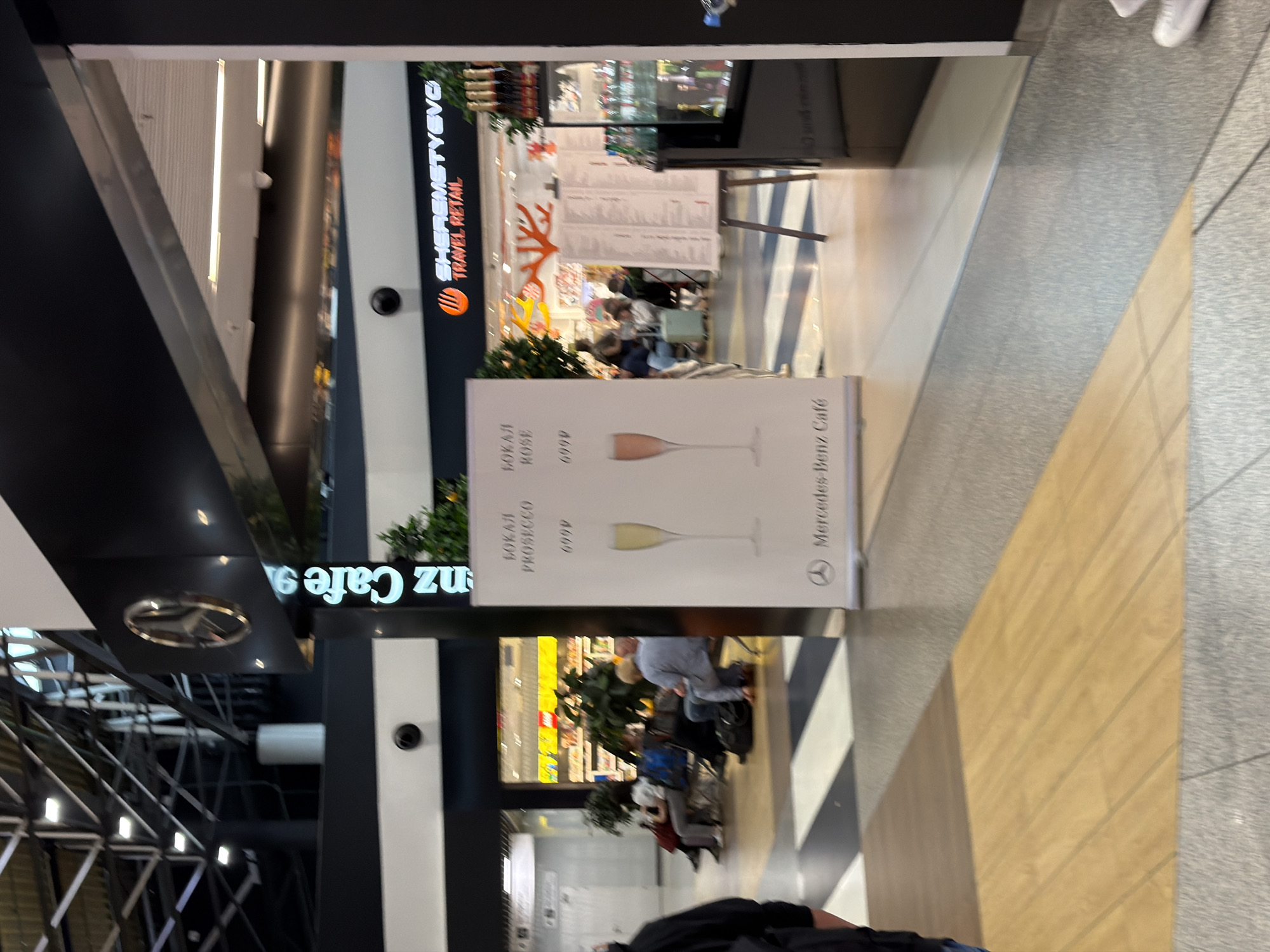 The image captures a view of the Mercedes-Benz Cafe at Sheremetyevo International Airport (SVO) in Perepechino, Russia. In the foreground, a poster displays two glasses of sparkling wine, one white labeled "PROSECCO" and one rose labeled "ROSE," with prices listed below. To the right of the poster, a display cabinet contains bottles of alcoholic beverages. In the background, several people are seated in a waiting area, with luggage nearby. The cafe's signage is visible above an entrance. The floor is a combination of wood-patterned tiles and grey stone tiles, with diagonal black and white stripes.