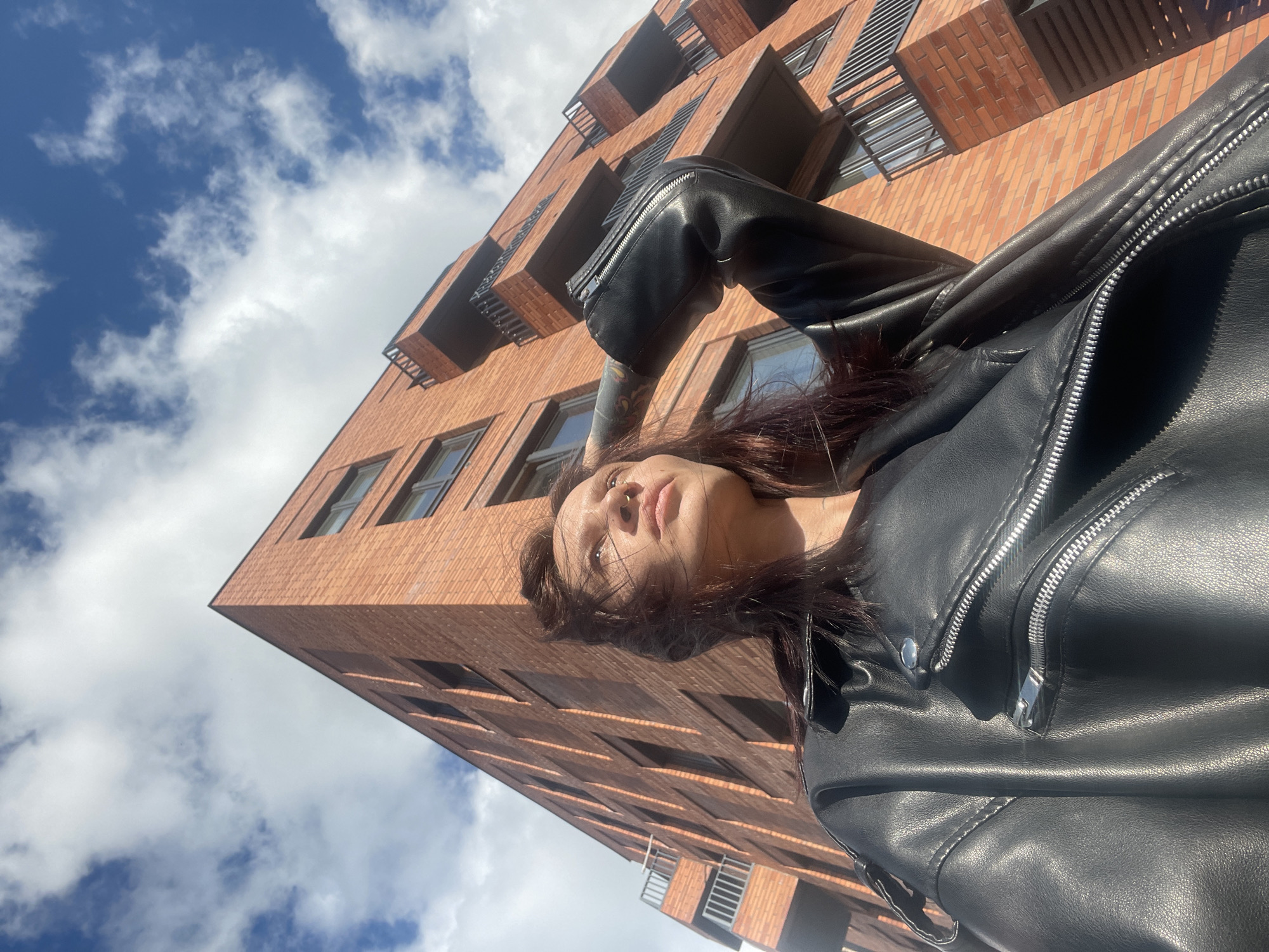 A low-angle, outdoor shot captures a person and a modern brick building against a bright blue sky. The main subject, positioned in the foreground and looking upwards, is a person with dark brown hair and a neutral expression. They are wearing a black leather jacket featuring prominent silver zippers and buttons. A septum nose piercing is visible, and what appears to be a tattooed arm is partially visible under the jacket sleeve.
In the background, a multi-story building made of reddish-brown bricks dominates the frame. It features numerous rectangular windows and dark-colored balconies or railings that project from its facade, indicating contemporary architecture. The extreme low-angle perspective emphasizes the height of the building, which appears to lean into the sky.
Above and around the building, the sky is clear blue with scattered white, fluffy clouds, suggesting a sunny day. The lighting is bright, casting some shadows and highlighting the textures of the brickwork and the leather jacket. The scene depicts an urban setting, with the provided location context being Izhevsk, Russia. No visible text is present in the image.