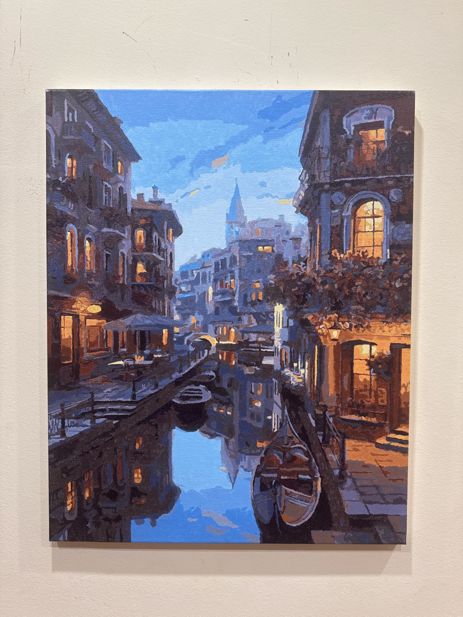The image is a painting of a canal scene in Venice, Italy, depicted at twilight or early evening. Tall, dark buildings with glowing windows line the canal, creating a sense of depth and atmosphere. The water reflects the warm light from the windows and the blue of the sky, adding to the serene mood. A gondola is partially submerged in the water, its dark form contrasting with the bright reflections. The architectural style of the buildings suggests a European city, likely Venice, with its characteristic canals and narrow waterways. The painting is done in a semi-realistic style, with visible brushstrokes contributing to its artistic quality. The image does not depict Hrodna, Belarus.