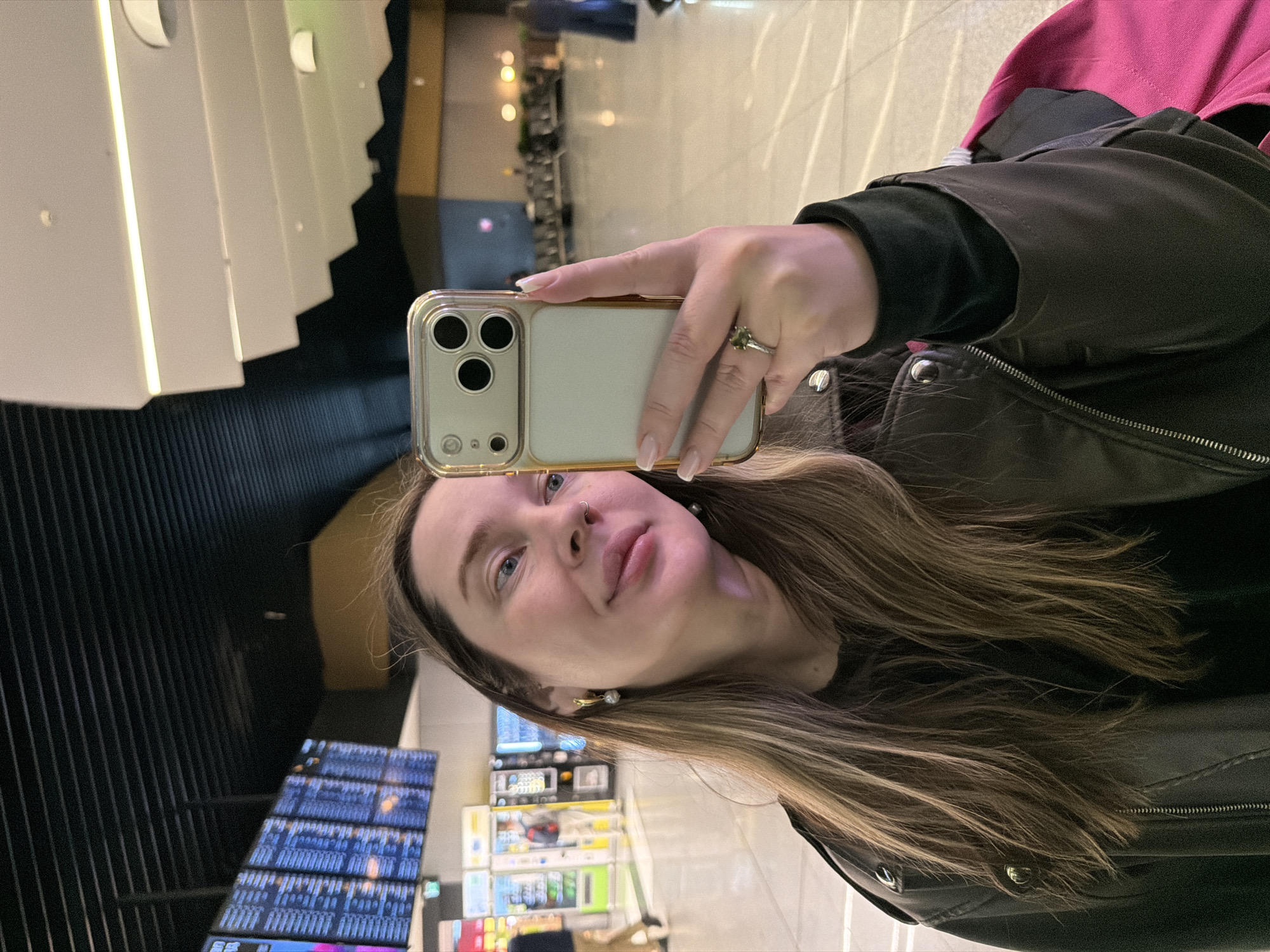 A person is taking a selfie in an airport in Khimki, Russia. The woman is holding a phone up to take a picture of herself. In the background, there are flight information screens. The person is wearing a jacket and has long brown hair.