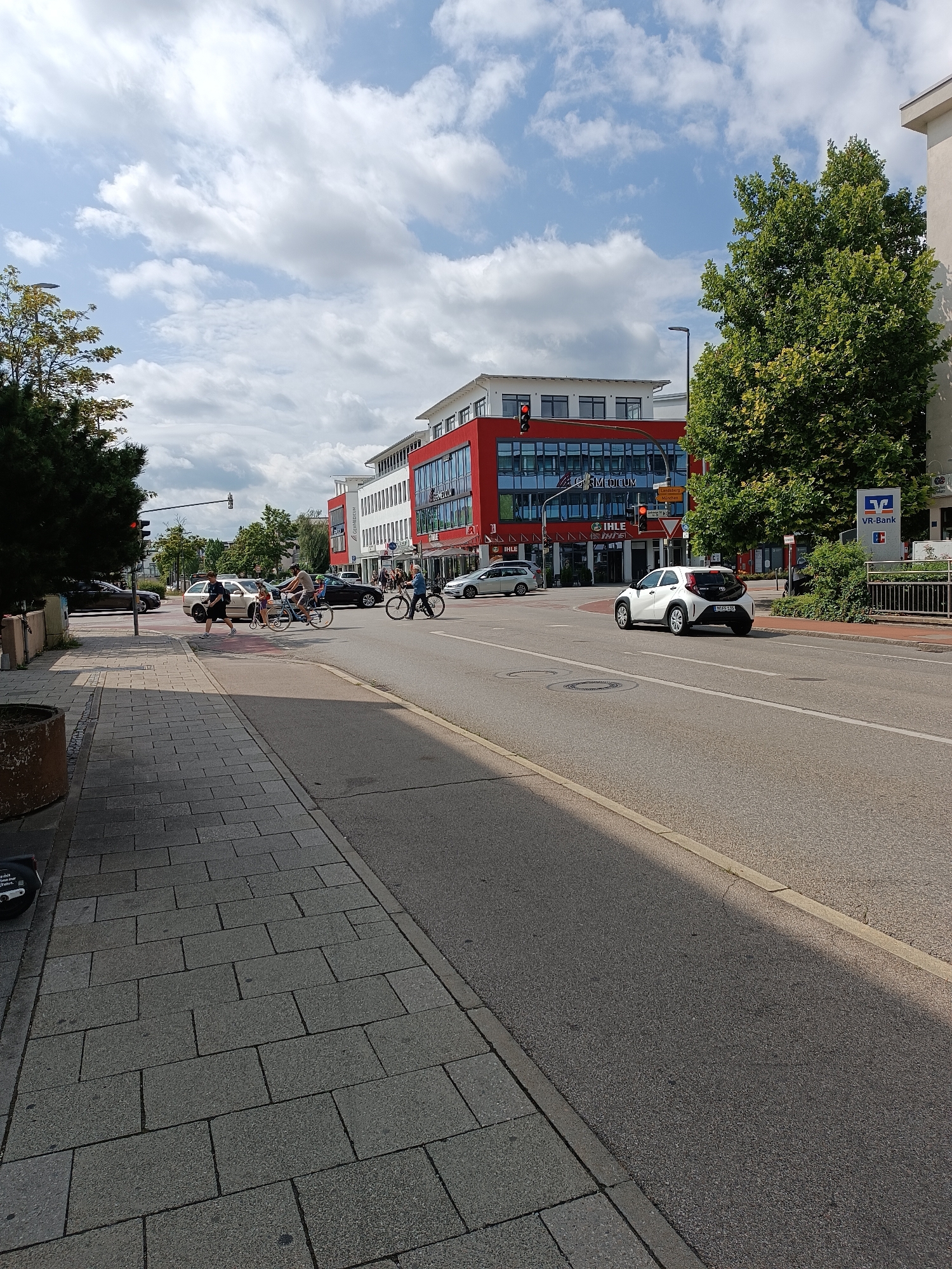 The image shows a street scene in Germering, Germany. A white car is driving on the right side of the road, and there is a building with a red facade on the left side of the street. Several people are on bicycles or walking on the sidewalk. The sky is cloudy with patches of blue.