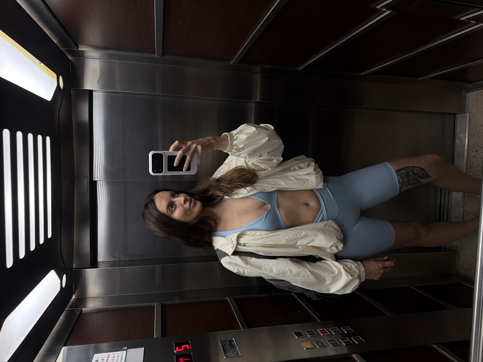 A woman is taking a selfie in an elevator. She is wearing a light blue sports bra and matching high-waisted biker shorts. An oversized, cream-colored jacket is open and draped over her shoulders. She has a backpack on her left shoulder. She is holding a white phone in her right hand, taking the picture in a mirror. The elevator walls are stainless steel with dark wood paneling. Above her are fluorescent lights. To her left is an elevator control panel with buttons and a digital display showing the number "5." The woman has long, dark brown hair and is looking slightly to her right. A tattoo is visible on her left thigh. The elevator floor appears to be concrete. The location is identified as Budva, Montenegro.