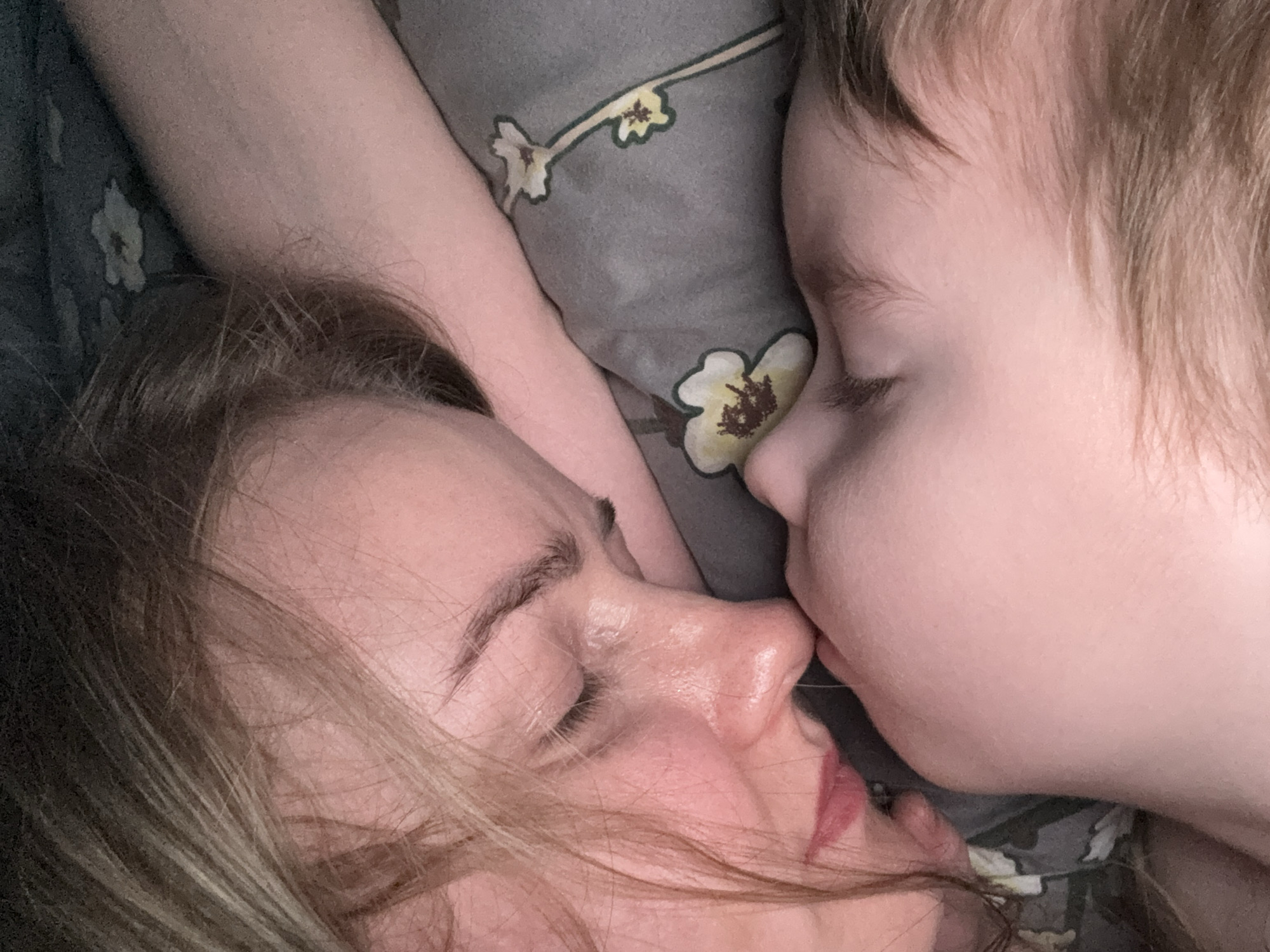 A close-up, intimate photograph captures two individuals, an adult female and a young child, resting together. The adult is positioned on the left, with light brown hair framing her face. Her eyes are closed, and her lips are slightly parted, revealing a hint of lipstick. Her skin appears soft and moisturized. The child, with fair skin and fine, light-colored hair, is on the right, also with closed eyes. Their cheek and forehead are pressed gently against the adult's face, suggesting a moment of tender affection, possibly a kiss or a snuggle.
Between and behind the subjects, a patterned fabric, likely a pillowcase or bedding, is visible. It features a muted grey-green background adorned with light yellow or cream-colored flowers that have dark brown centers and delicate green stems. The composition focuses tightly on the faces, emphasizing their connection. The soft, natural lighting contributes to a serene and peaceful atmosphere. The scene portrays a quiet, loving moment of rest or sleep. No visible text appears in the image. The location context for this tender moment is Chaikovsky, Russia.